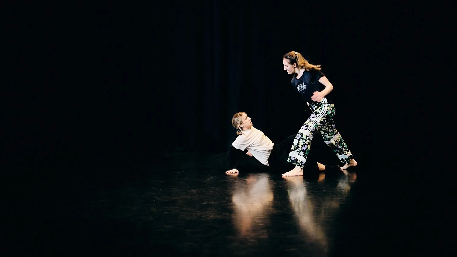 Two women practicing a dance or performance move on a dark stage, with one woman on the floor and the other standing over her.