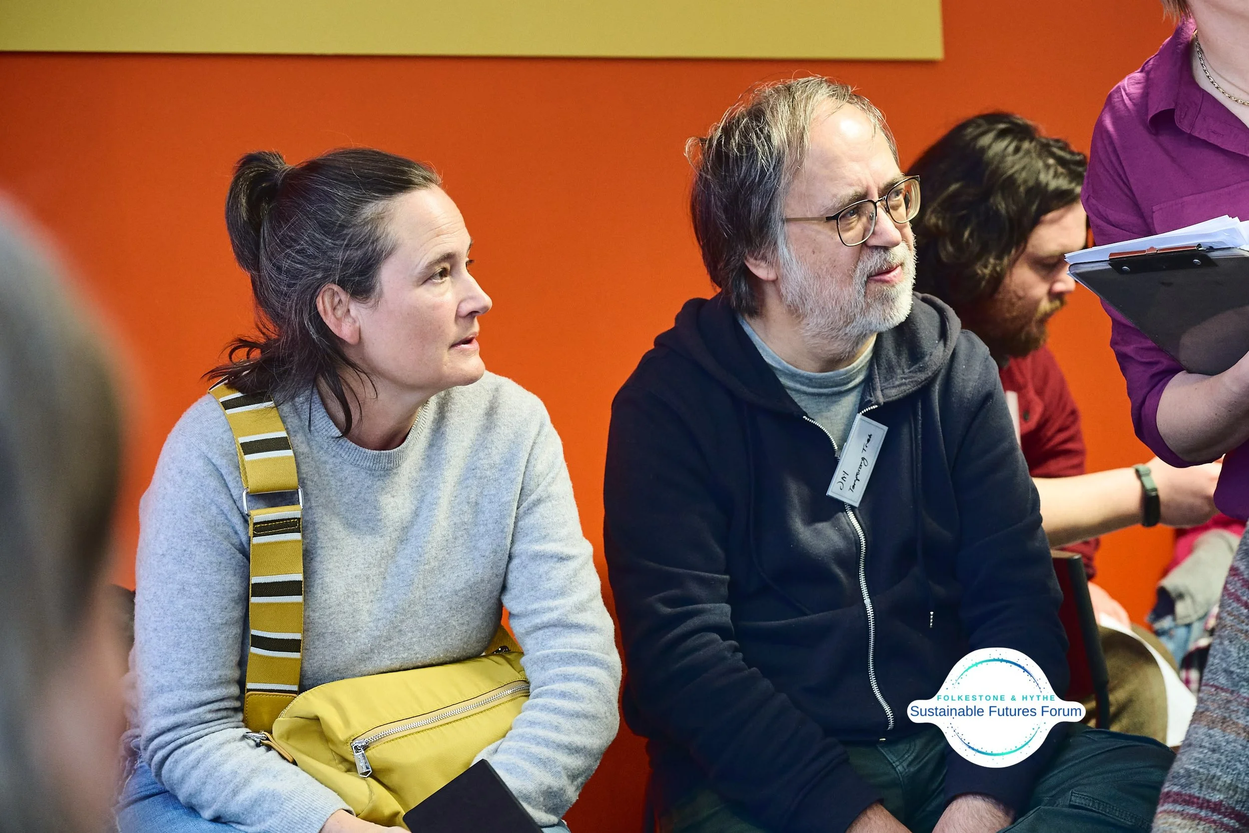 Two people sitting in a row at a forum, listening attentively. A woman with dark hair tied back wearing a light gray sweater and mustard yellow bag, and a man with glasses and gray hair in a black hoodie. An orange wall in the background with a logo 