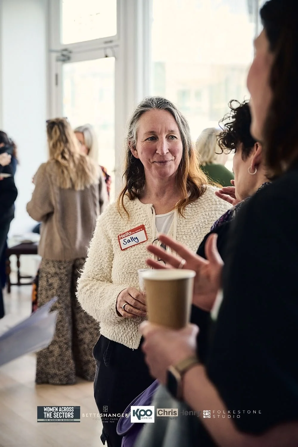 A woman with long gray hair, wearing a cream-colored sweater, holding a coffee cup, and a name tag reading 'Sally', is talking to another woman at a networking event. Background shows other women engaged in conversations near large windows.