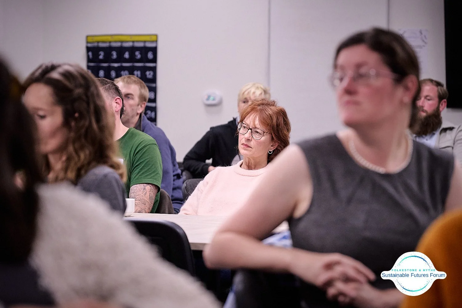People attending a forum or seminar, sitting at tables and listening attentively, with a visible sign on the wall that reads 'Sustainable Futures Forum'.