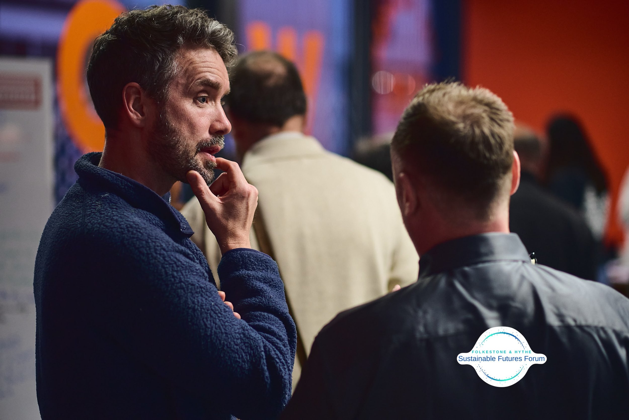 Two men are engaged in conversation at a public event, with one facing the camera in profile and the other with his back to the camera. The background is blurred, featuring a colorful signage and several people.