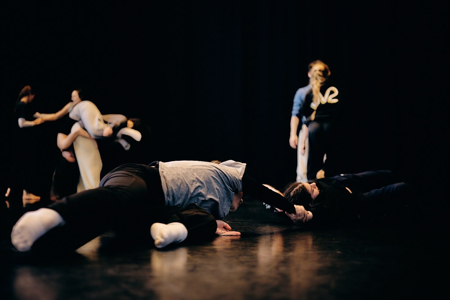 Performers on stage, some lying on the floor and others standing, with a dark background. Two performers in animal costumes are visible among the group.