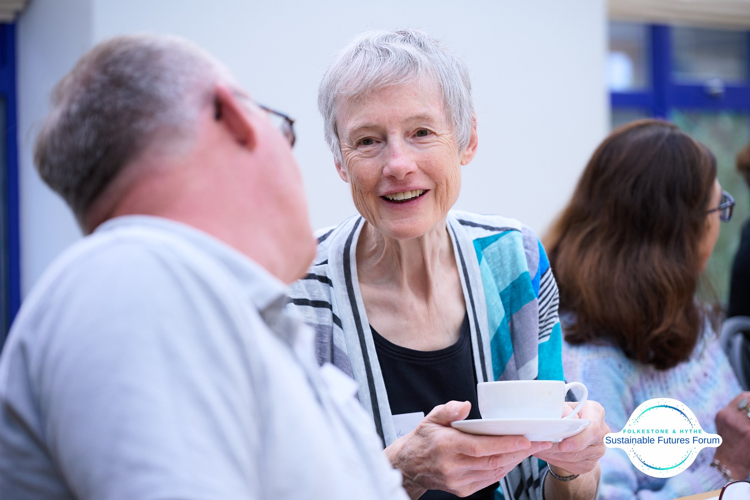 An elderly woman with short gray hair smiling and holding a cup and saucer, engaged in conversation with a man with glasses and gray hair, at a social gathering.
