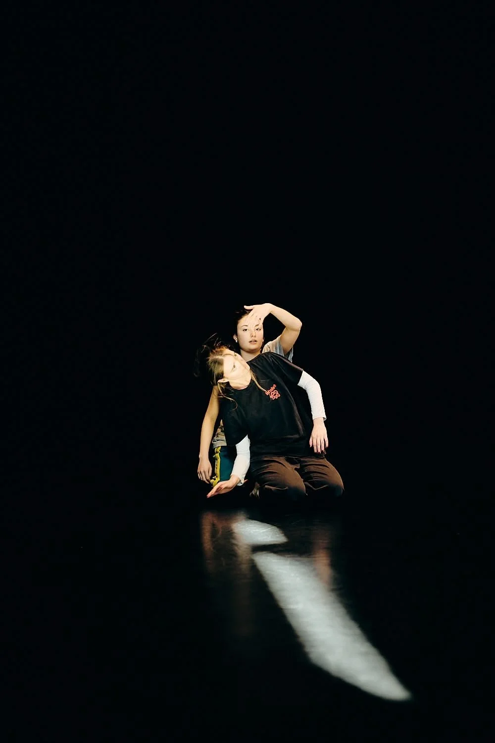 Two dancers performing on a dark stage with a bright white light reflection on the floor, one sitting and the other leaning over, with their heads touching.