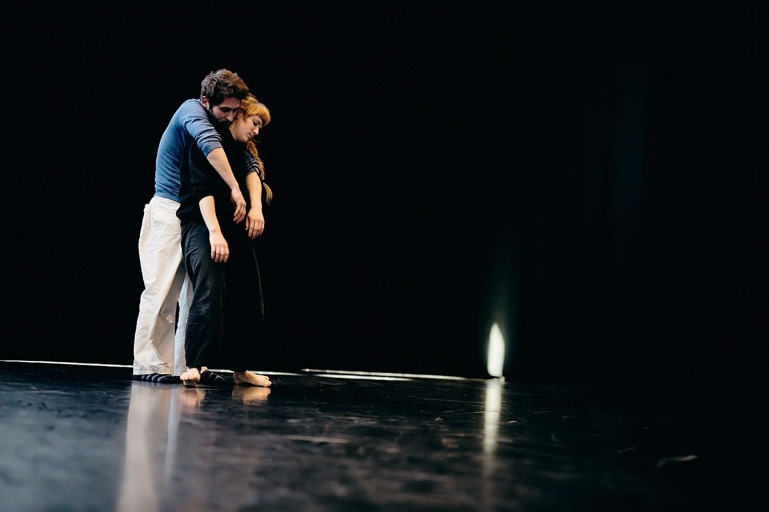 Two people stand closely together on a dark stage, their heads leaning against each other, in an expression of support or connection.