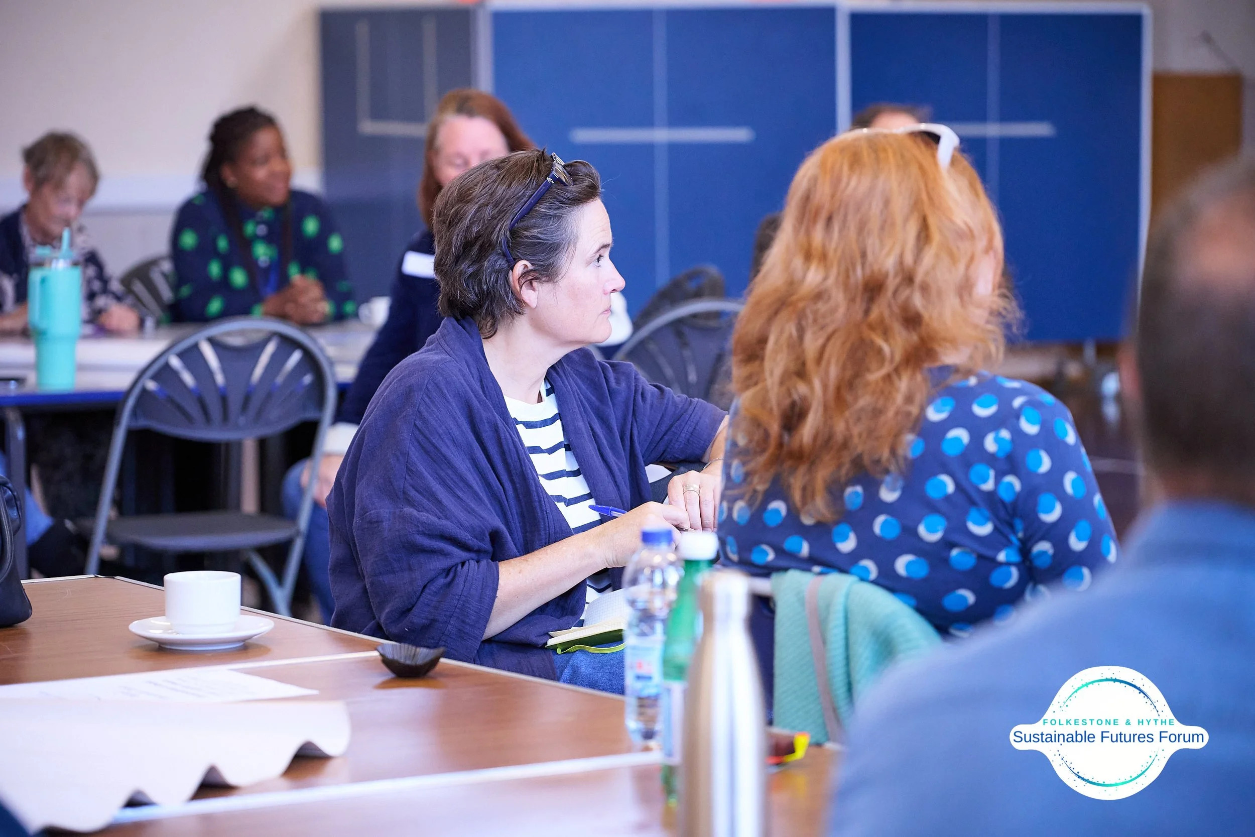 Group of people attending a Sustainable Futures Forum at Folkesstone & Hythe, sitting at tables with notebooks, water bottles, and cups.
