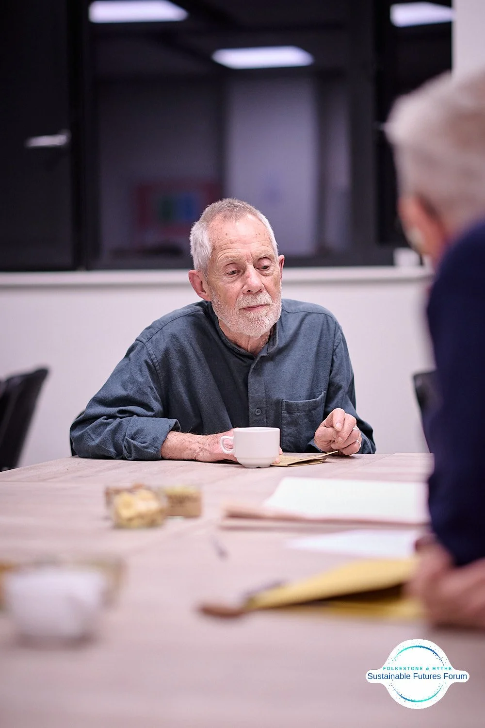 An elderly man with gray hair and a beard sitting at a conference table with a cup and documents, listening to another person. There are papers and a small snack on the table, and a logo in the bottom right corner.