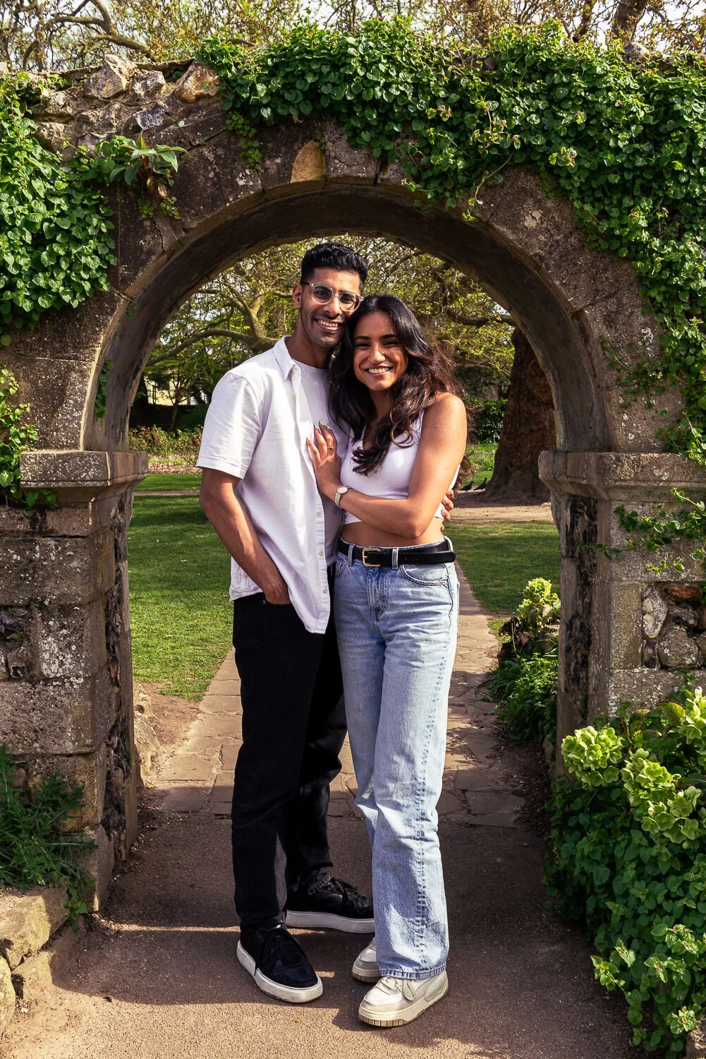 A young couple smiling and embracing in front of an arched stone gateway covered in green ivy in a garden with trees and green grass.