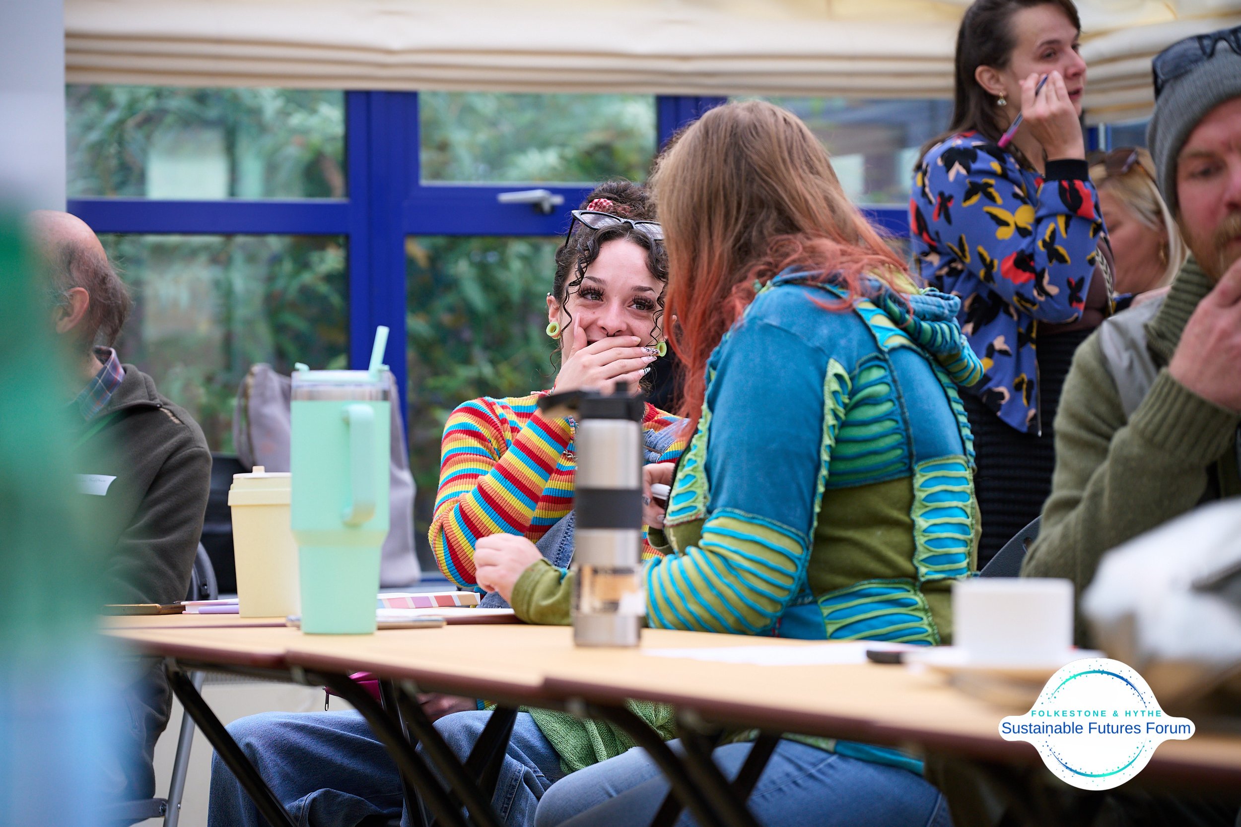 Group of people sitting at a table, engaged in conversation, with some young women laughing and smiling, in an indoor setting with large windows showing greenery outside.
