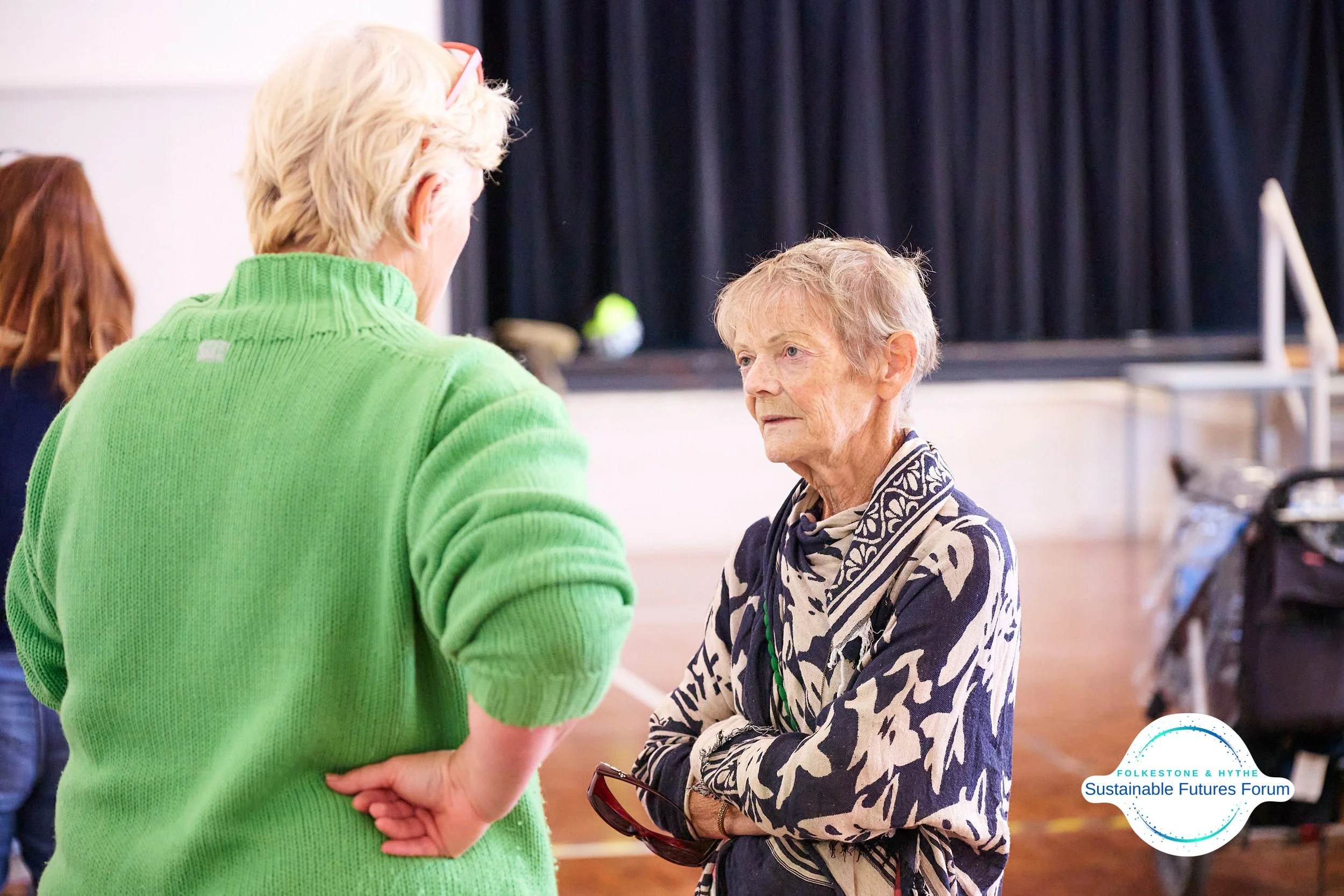 Two women engaged in a conversation at an indoor event, one wearing a bright green sweater and the other wearing a black and white patterned jacket, with a black curtain and a wheelchair visible in the background.