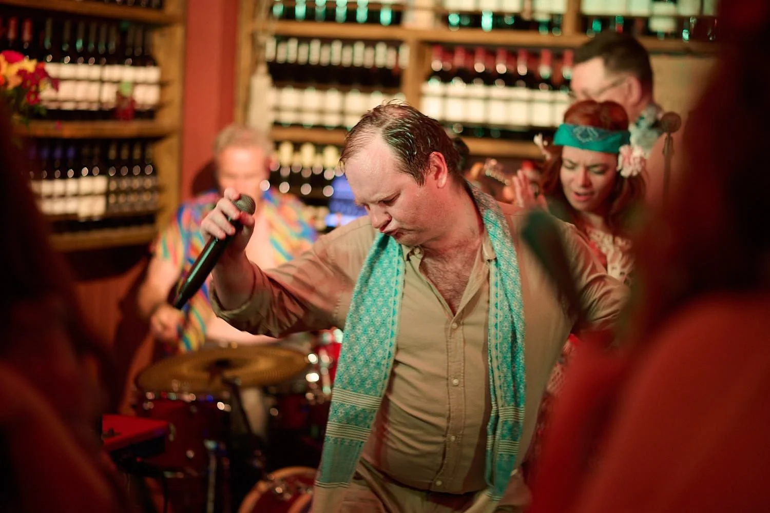 A man with a scarf around his neck, holding a microphone and dancing at a lively party with other people in the background.