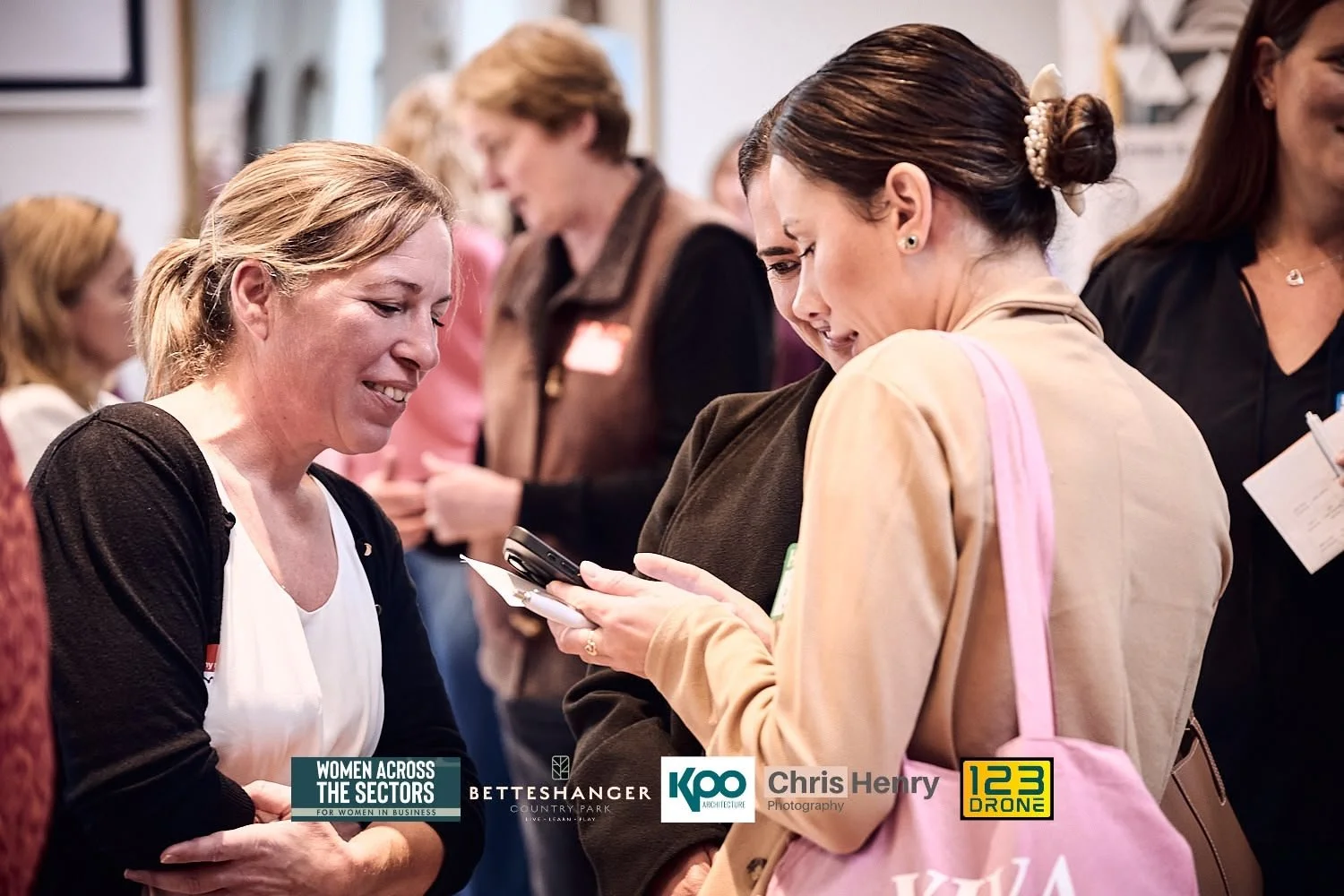 Two women smiling and looking at a smartphone at a networking event, with other women in the background.