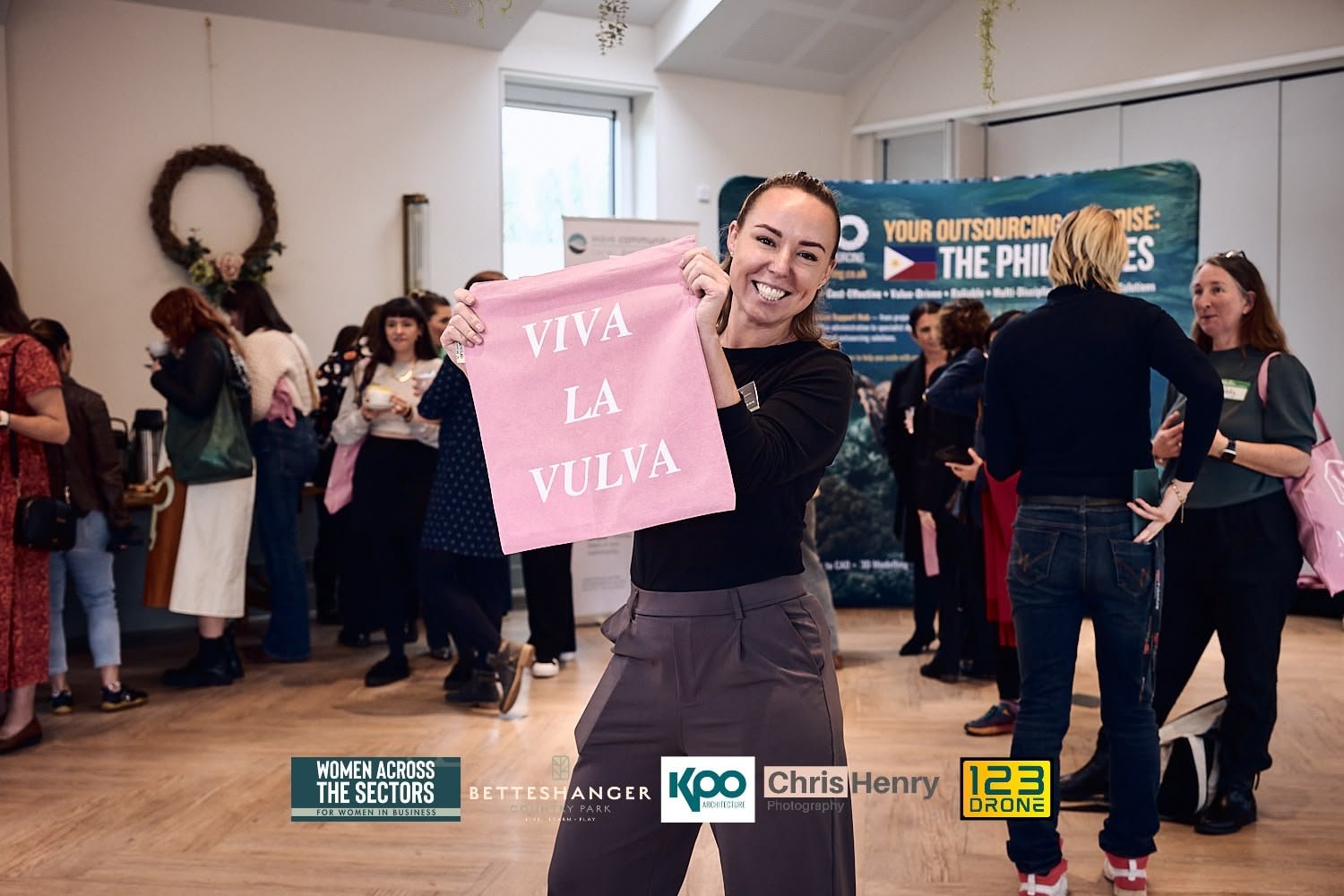 A woman with brown hair smiling and holding a pink sign that says "VIVA LA VULVA" at a gathering or event with multiple people in the background.
