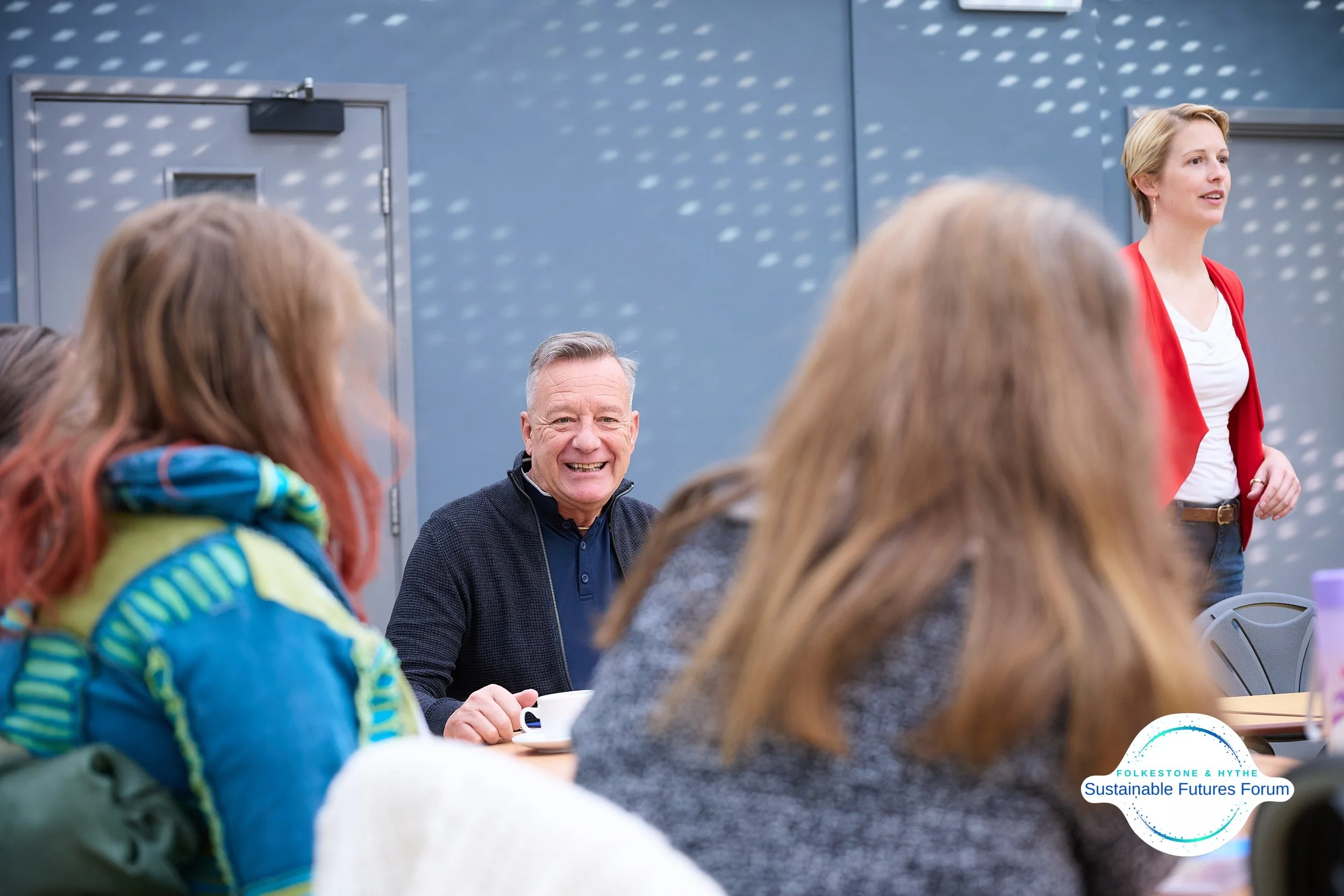 A woman in a red blazer speaking to a group during a forum, with a man smiling and holding a coffee cup at a table. The forum is part of the Folkesetone & Hythe Sustainable Futures Forum.