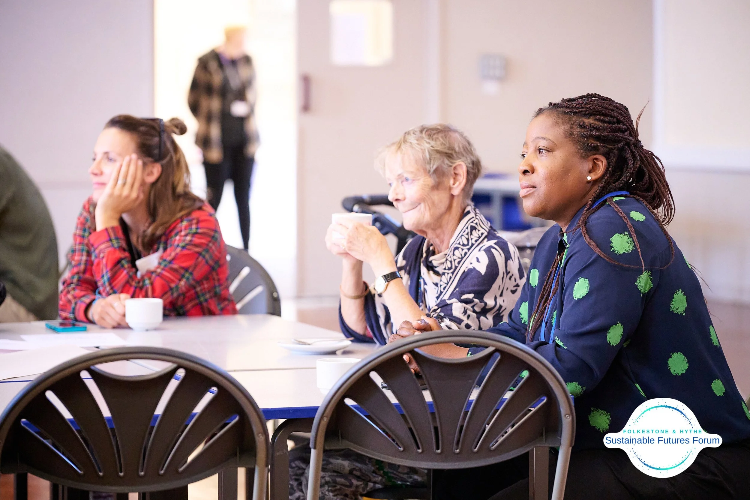 Three women sitting at a table during a forum, with a woman in the background standing near a door, in a well-lit room. The women are engaged and listening attentively.