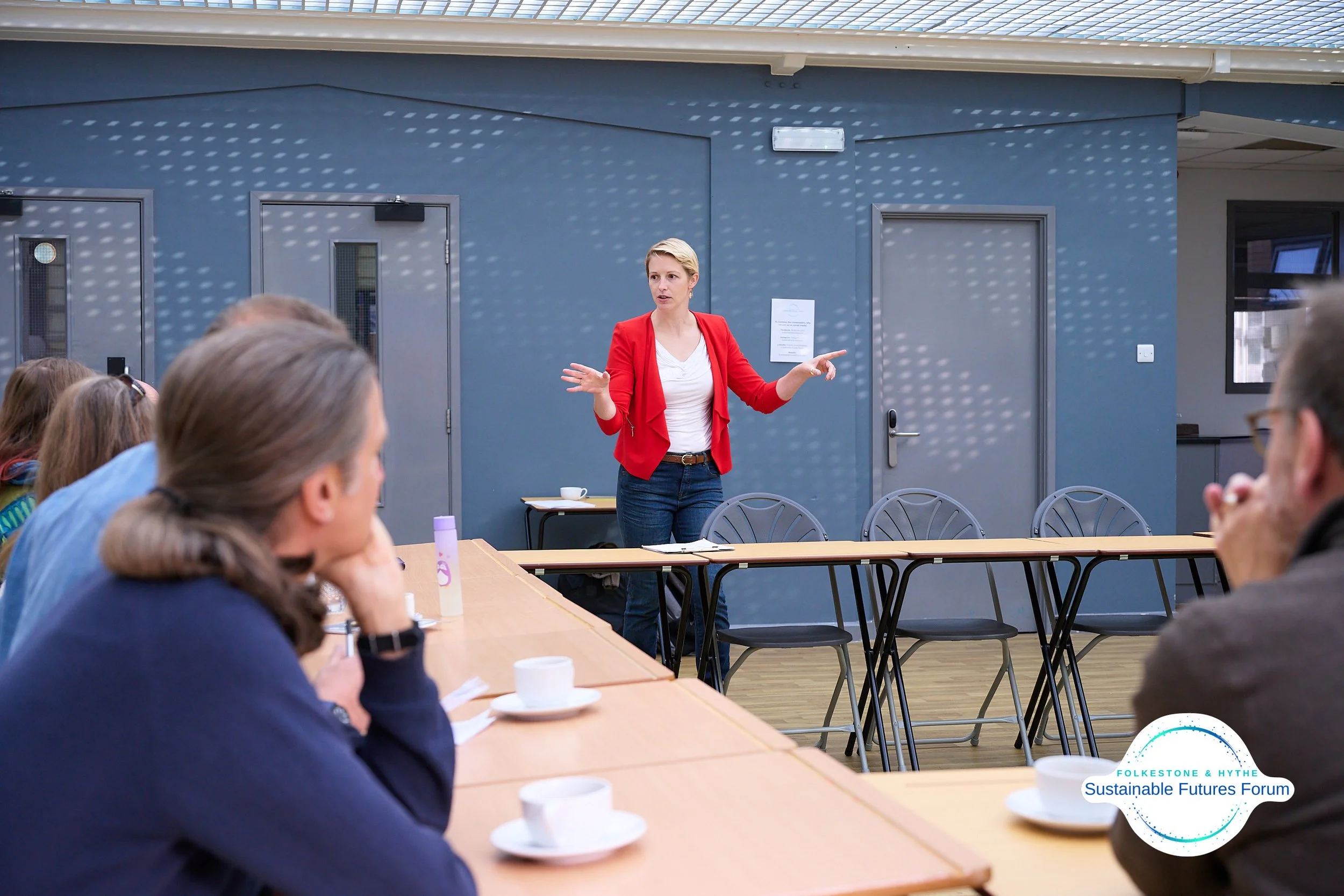 A woman in a red blazer is giving a presentation to a group of people seated at a table in a conference room. The room has blue walls and large windows with sunlight creating patterns on the walls.