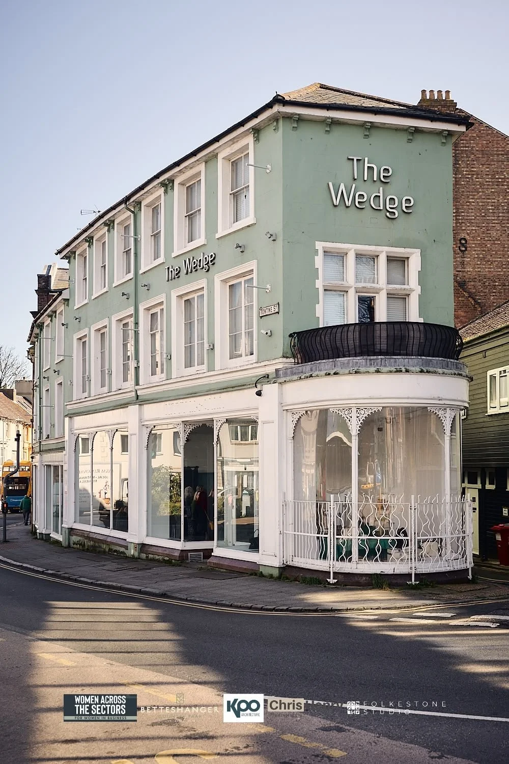 A three-story building painted green with white trim, named "The Wedge," with large windows and a rounded balcony on the second floor. It is located on a street corner.