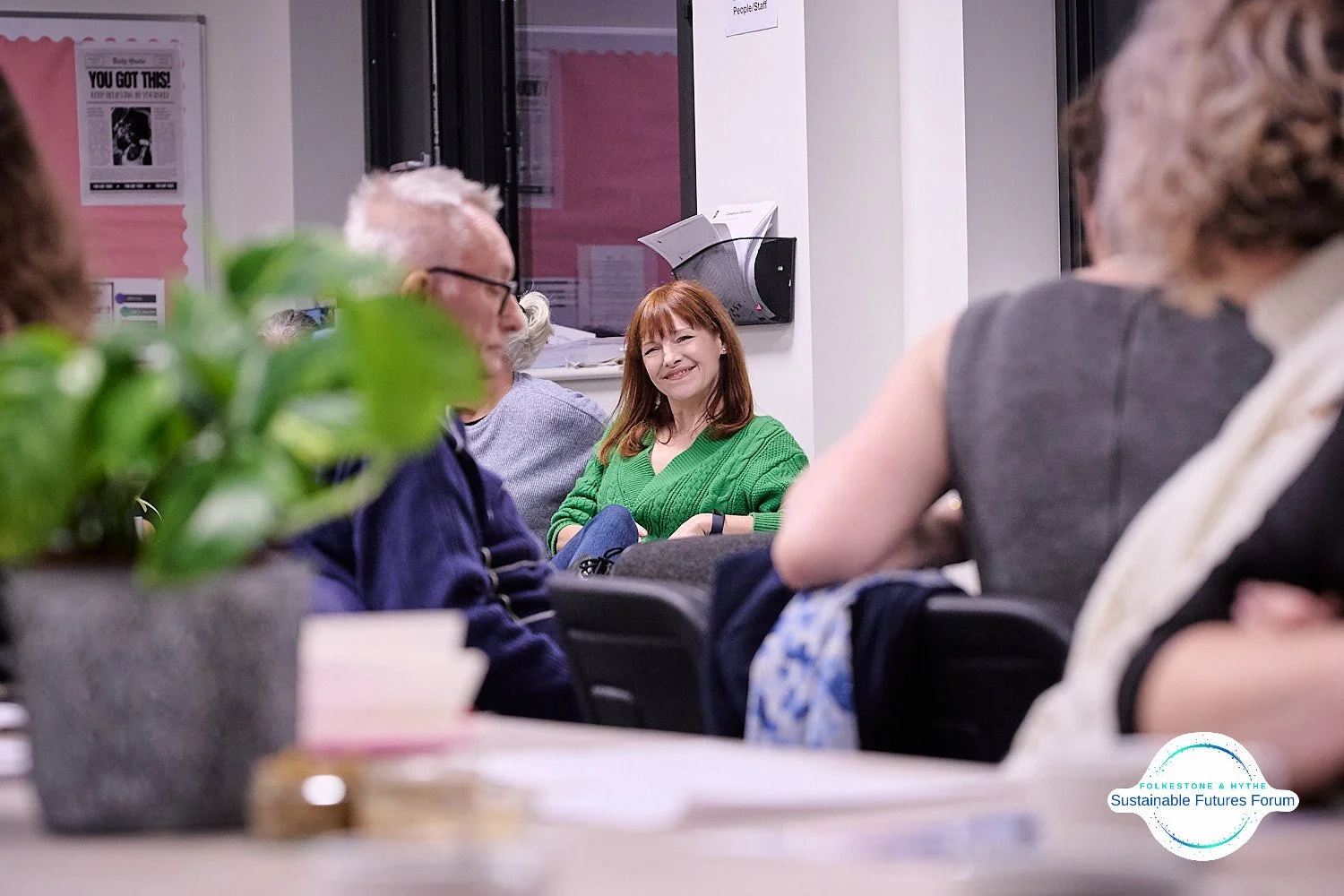 A woman in a green sweater smiling during a meeting, with other attendees seated around her in a conference room.