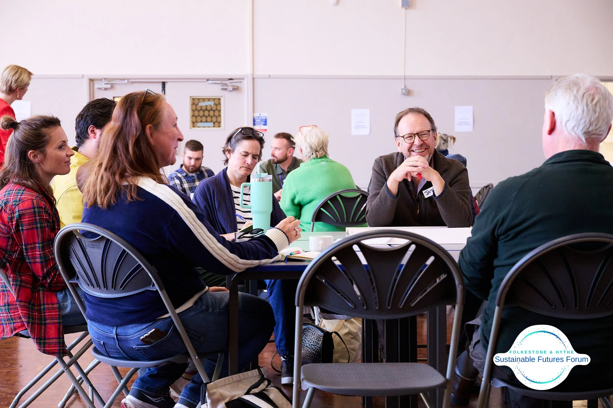 A group of people sitting around tables, engaging in a discussion at the Sustainable Futures Forum by Folkestone & Hythe, with a man smiling and talking to a woman and others listening attentively.