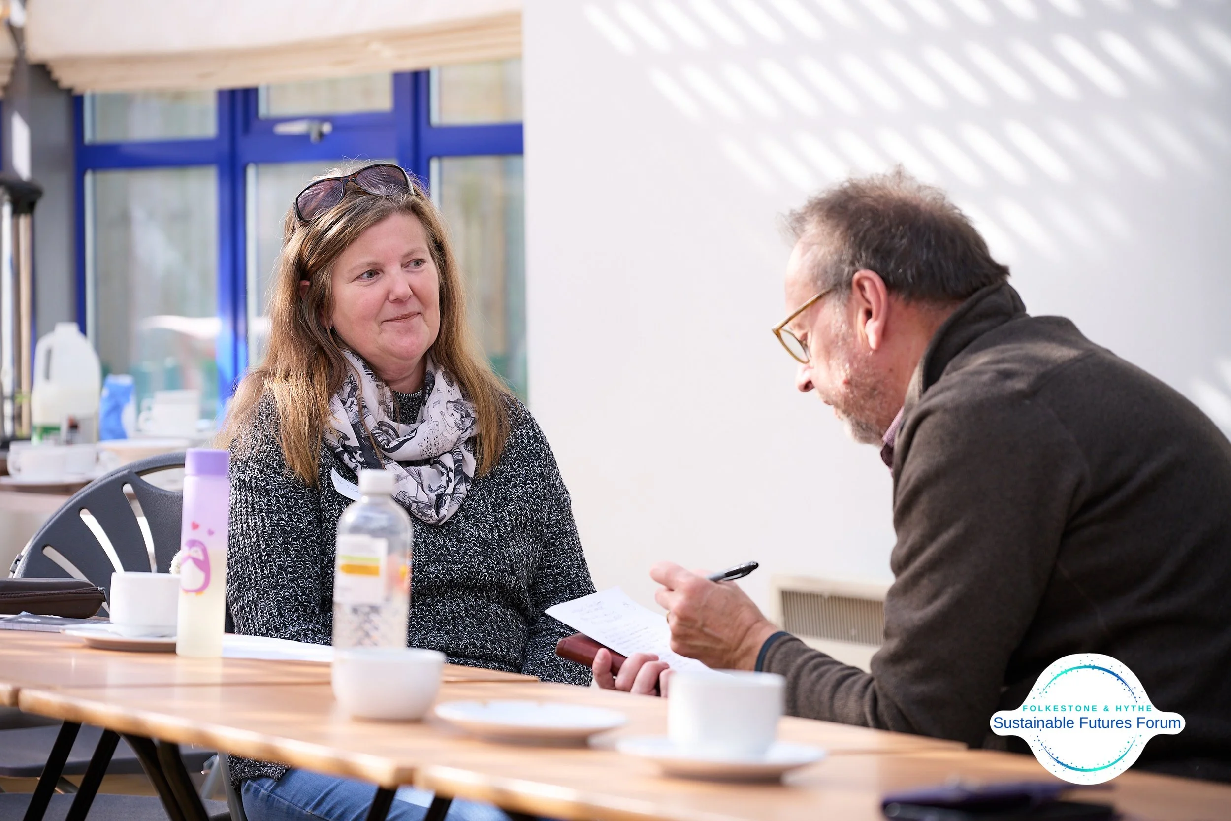 A woman and a man sitting at a wooden table in a bright room, engaging in a conversation or an interview, with the woman wearing a patterned scarf and the man focusing on a notepad or phone.