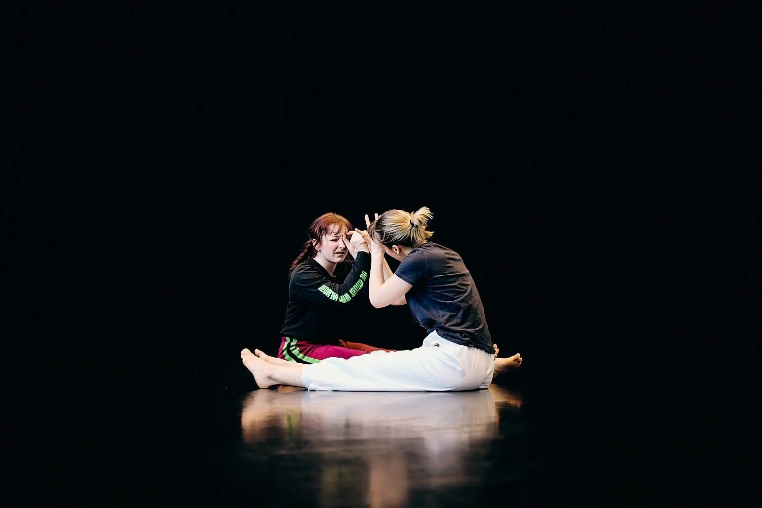 Two women practicing Brazilian Jiu-Jitsu on a black mat, with black background, one in a black sweatshirt and green-striped pants, the other in a black t-shirt and white pants, engaged in a close grip.