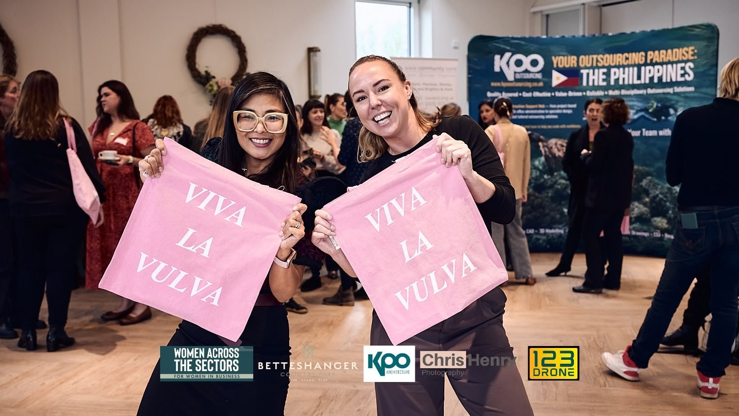 Two women smiling and holding pink cloth signs that say 'VIVA LA VULVA' at a women-focused event with a crowd in the background and a banner about the Philippines.