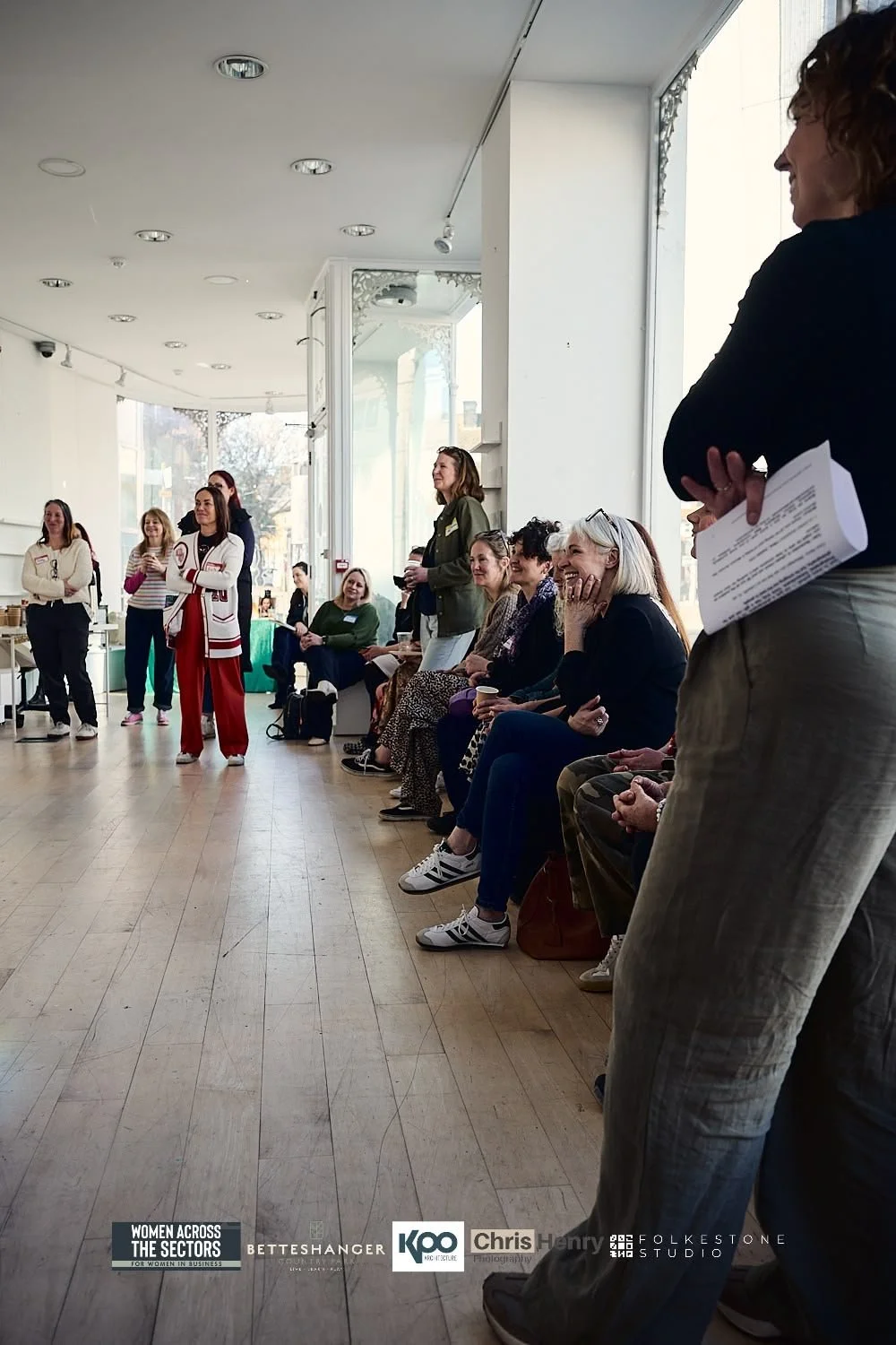 Women gathered in a bright indoor space, some sitting and some standing, engaged in a discussion or presentation, with smiles and attentive expressions.