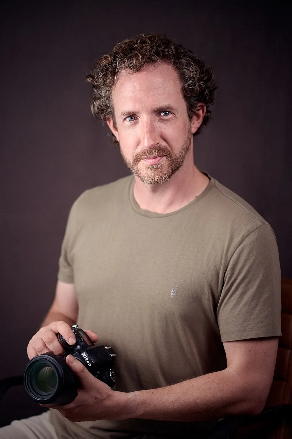Portrait of a man with curly brown hair, blue eyes, and a beard, wearing a brown t-shirt against a brownish background.