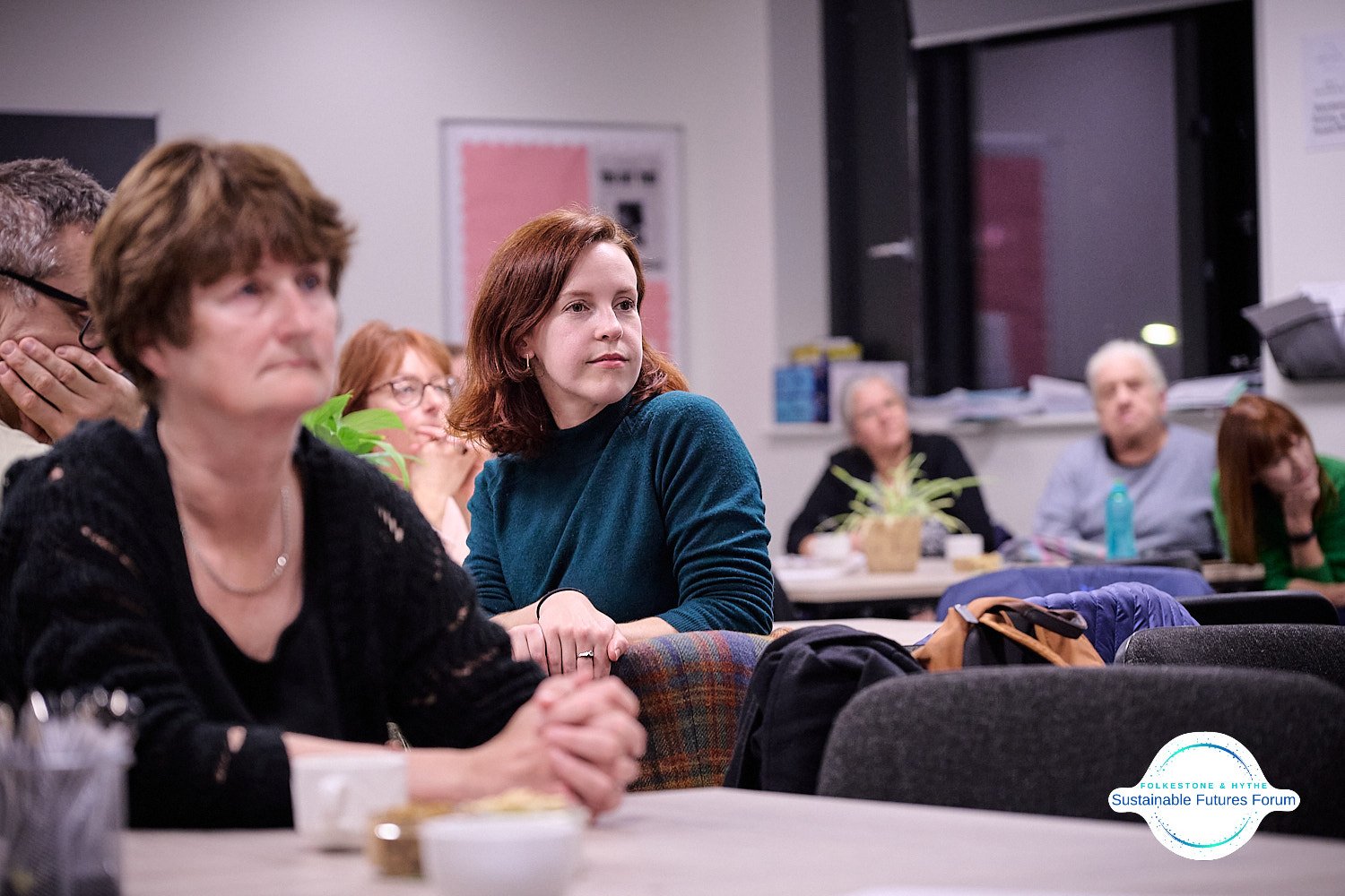 Group of people attending a meeting in a conference room, some listening attentively and others resting or thinking, with a logo for the Folkestone & Hythe Sustainable Futures Forum in the bottom right corner.