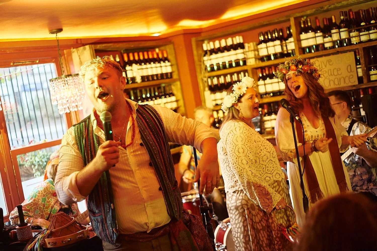 A man singing into a microphone at a lively gathering with women wearing floral crowns, inside a wine shop or bar with bottles on shelves.