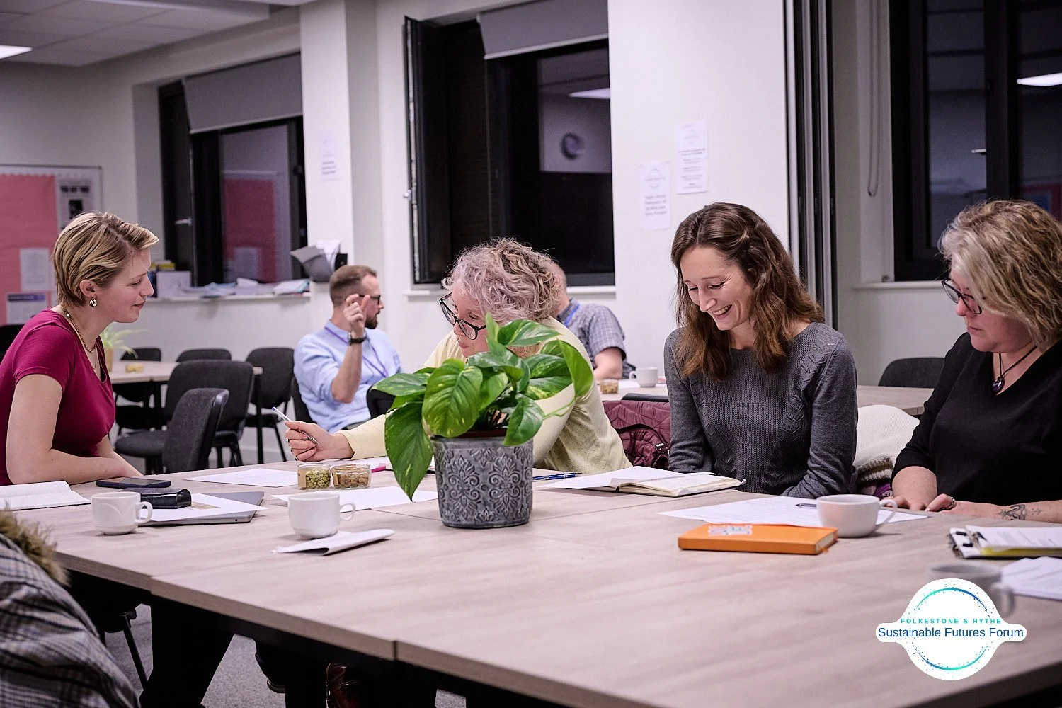 A group of five women sitting around a table in a meeting room, engaging in conversation, with notebooks, cups, and snacks on the table, and a potted plant in the center.