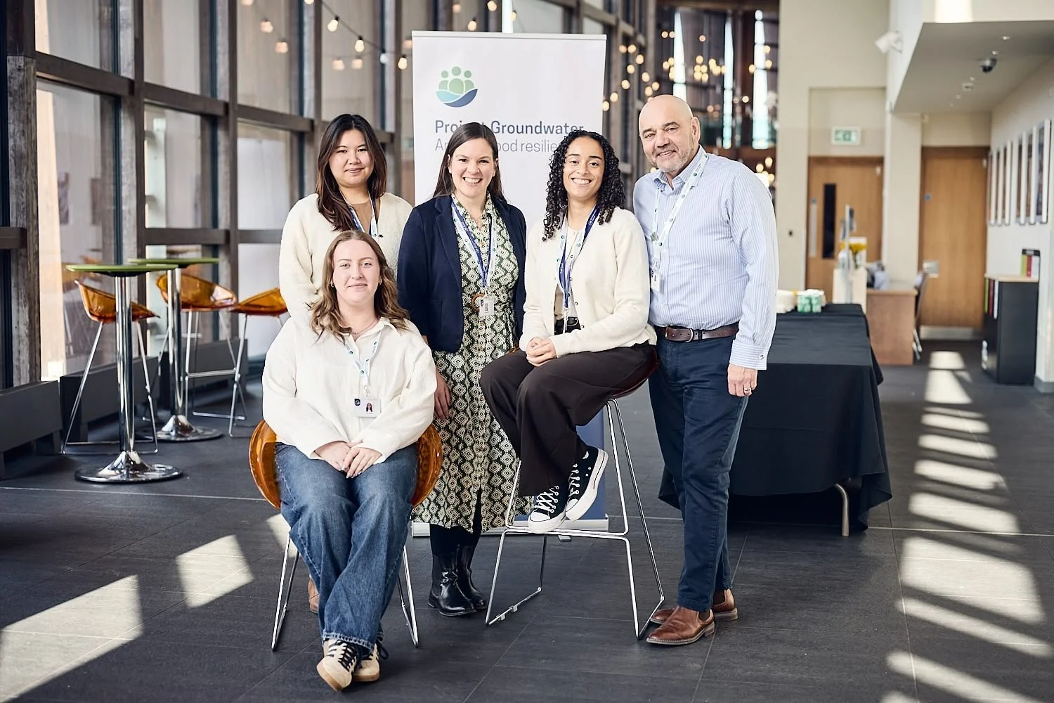 Group of six people posing inside a modern building with a banner in the background that reads "Protect Groundwater, Adapt to Food Resilience." The group includes five women and one man, all smiling, with some wearing conference lanyards.