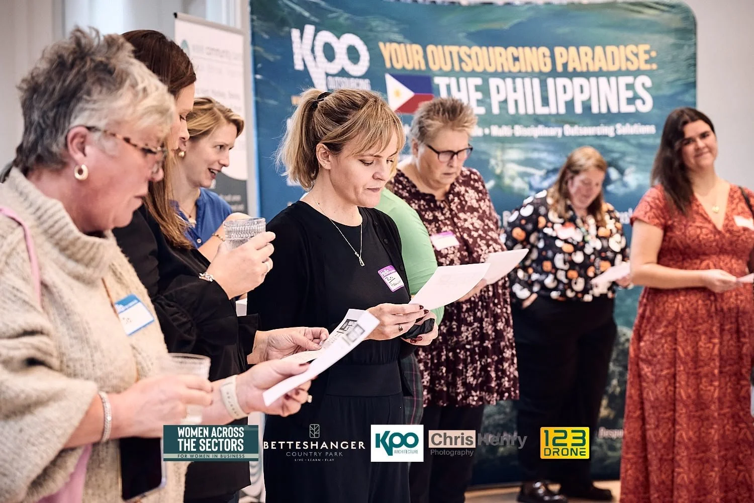 A group of women standing side by side, reading papers at a conference. A large banner in the background reads 'KOO Your Outsourcing Paradise: The Philippines' and features images of the Philippines flag and landscape. The women are holding glasses a