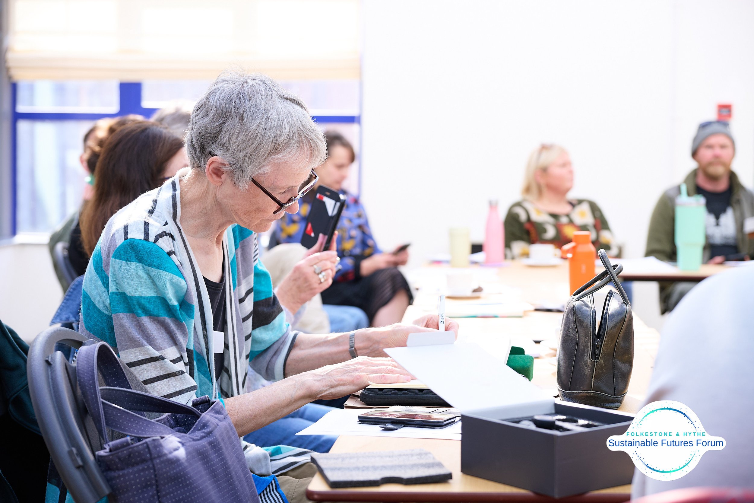 Older woman taking notes at a conference table during a sustainability forum, with other attendees using smartphones in the background.