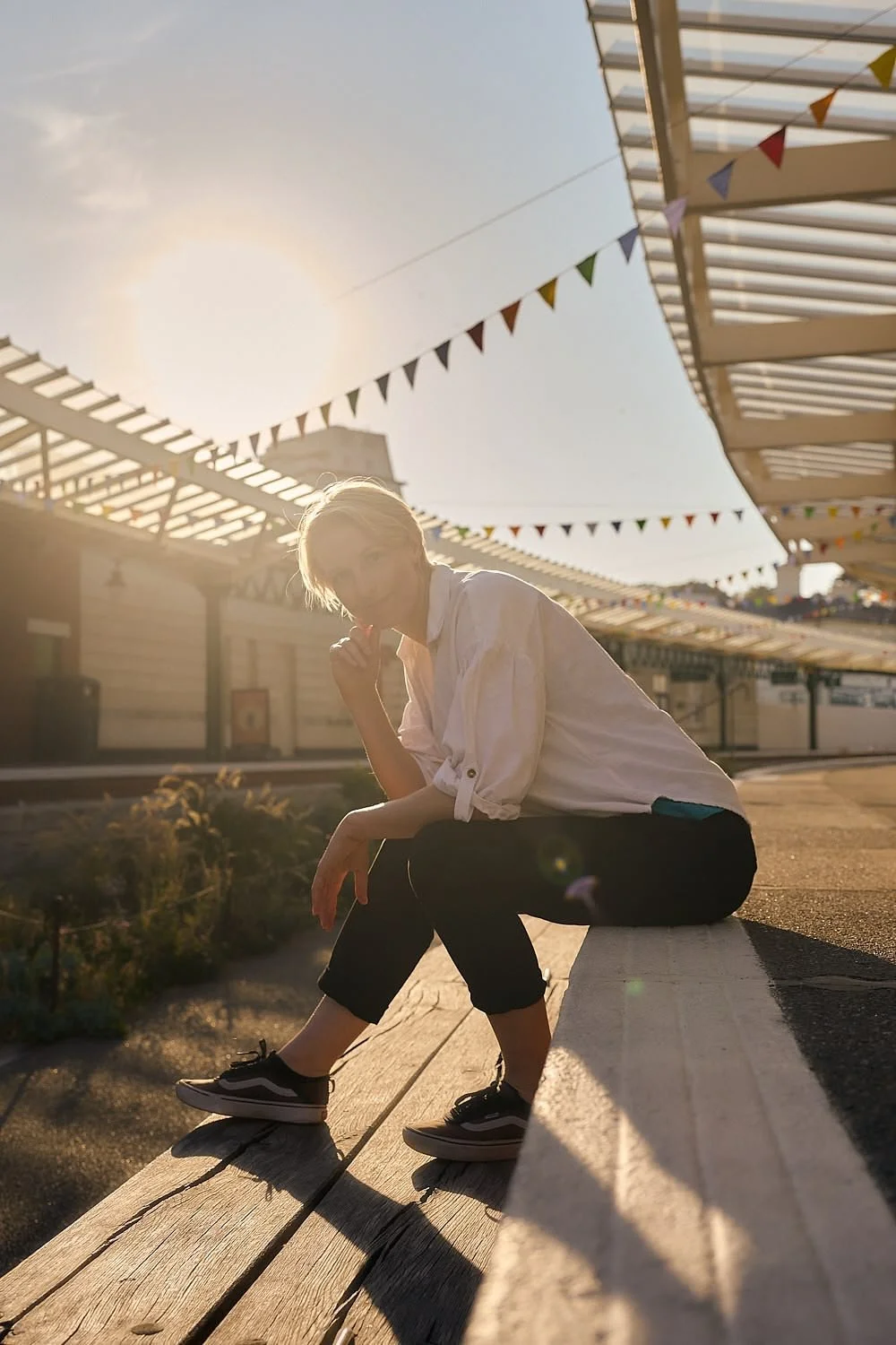 Person with short blonde hair in a white shirt and black pants sitting on a wooden ledge at an amusement park during sunset, with colorful pennant banners and modern light structures in the background.