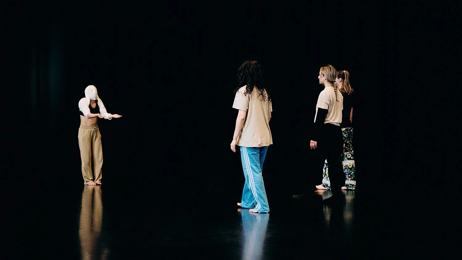 Four women with casual clothing and barefoot, standing on a dark stage, engaged in a dance or acting rehearsal.