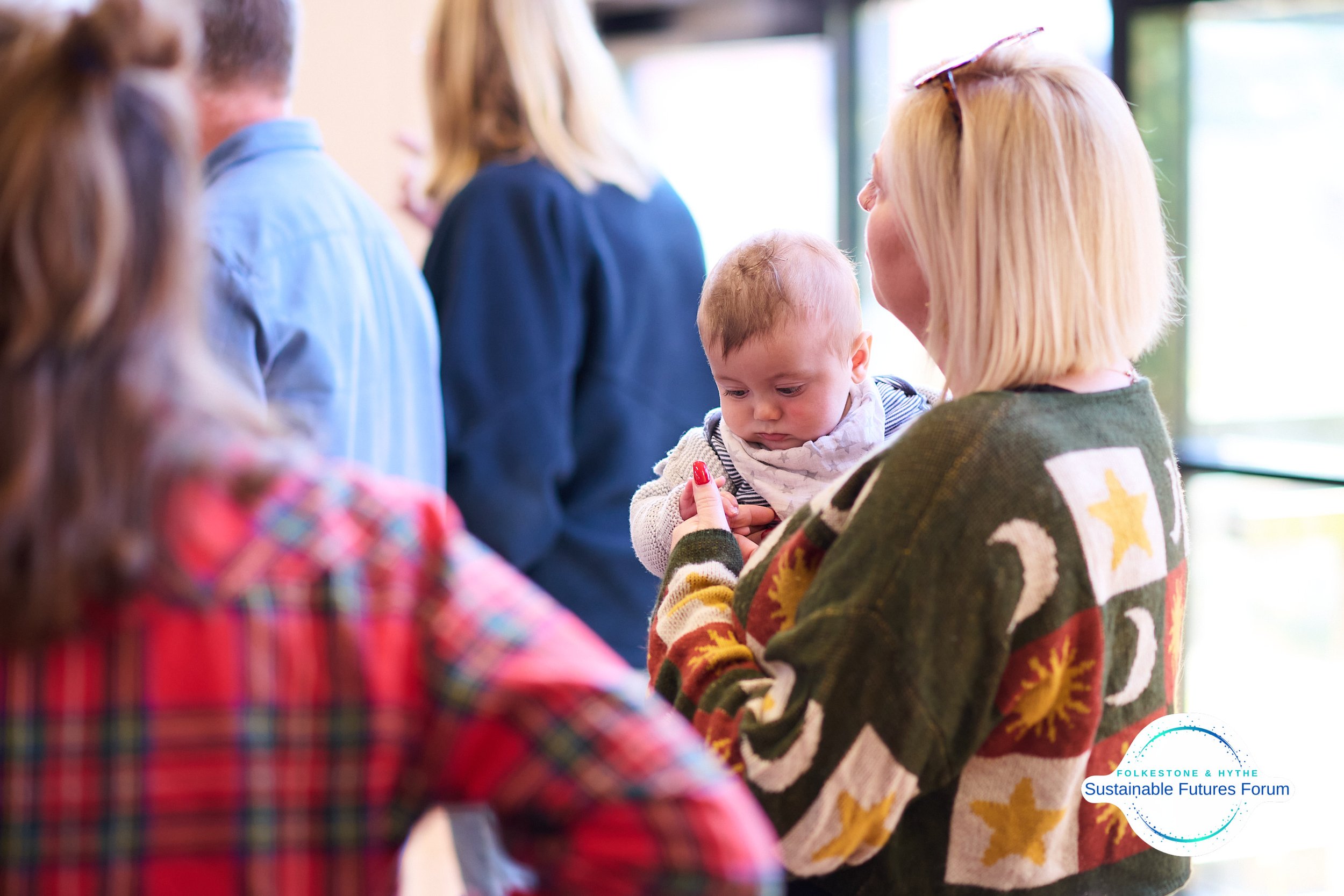 A woman with blonde hair wearing a colorful sweater holds a young child, who looks somewhat serious, in her arms. The scene appears to be a gathering of adults, some visible in the background, in a well-lit indoor space. The woman's sweater features 