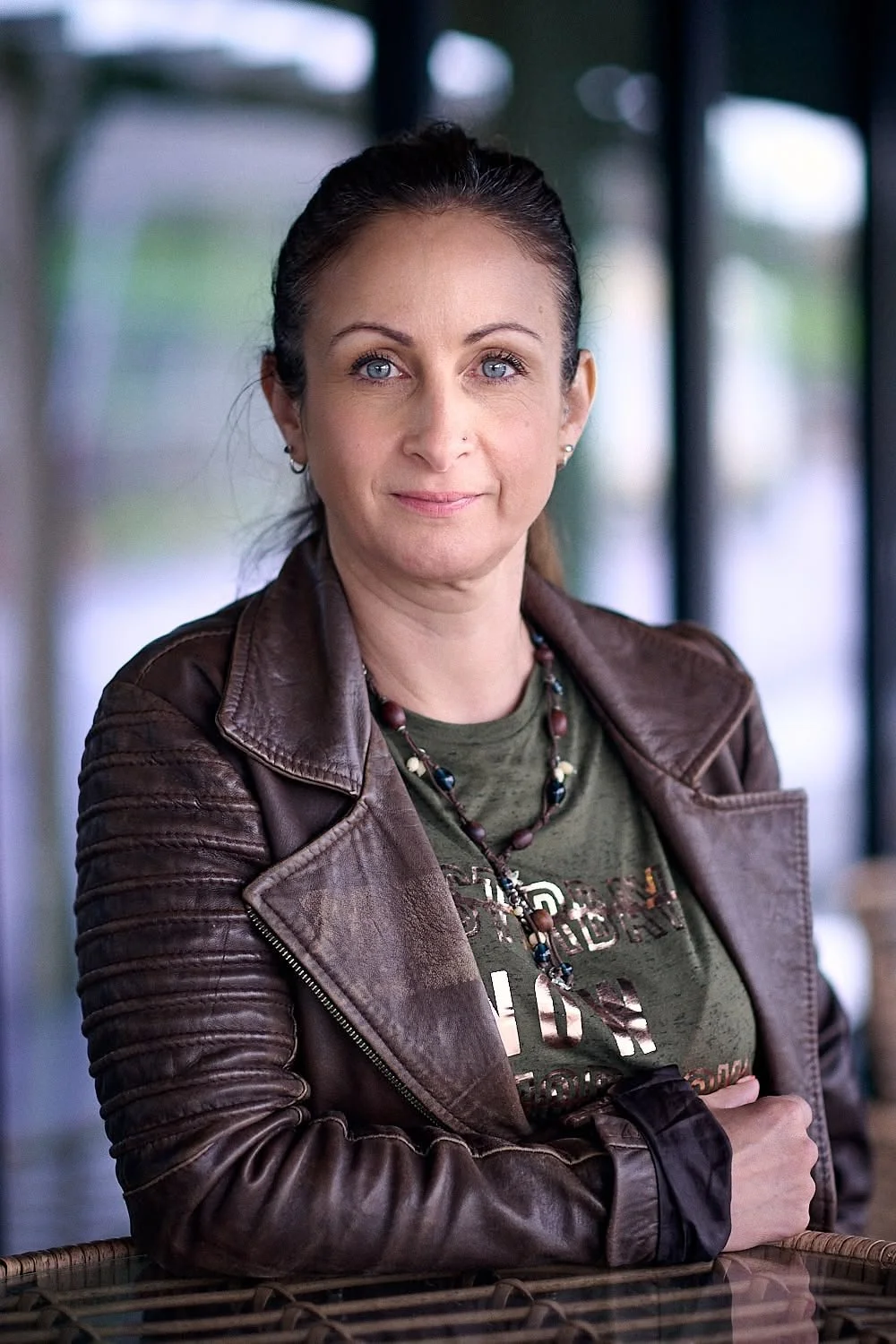 A woman with blue eyes and dark hair tied back, wearing a brown leather jacket over an olive green t-shirt with printed text, and a beaded necklace, standing indoors near a window with blurred background.