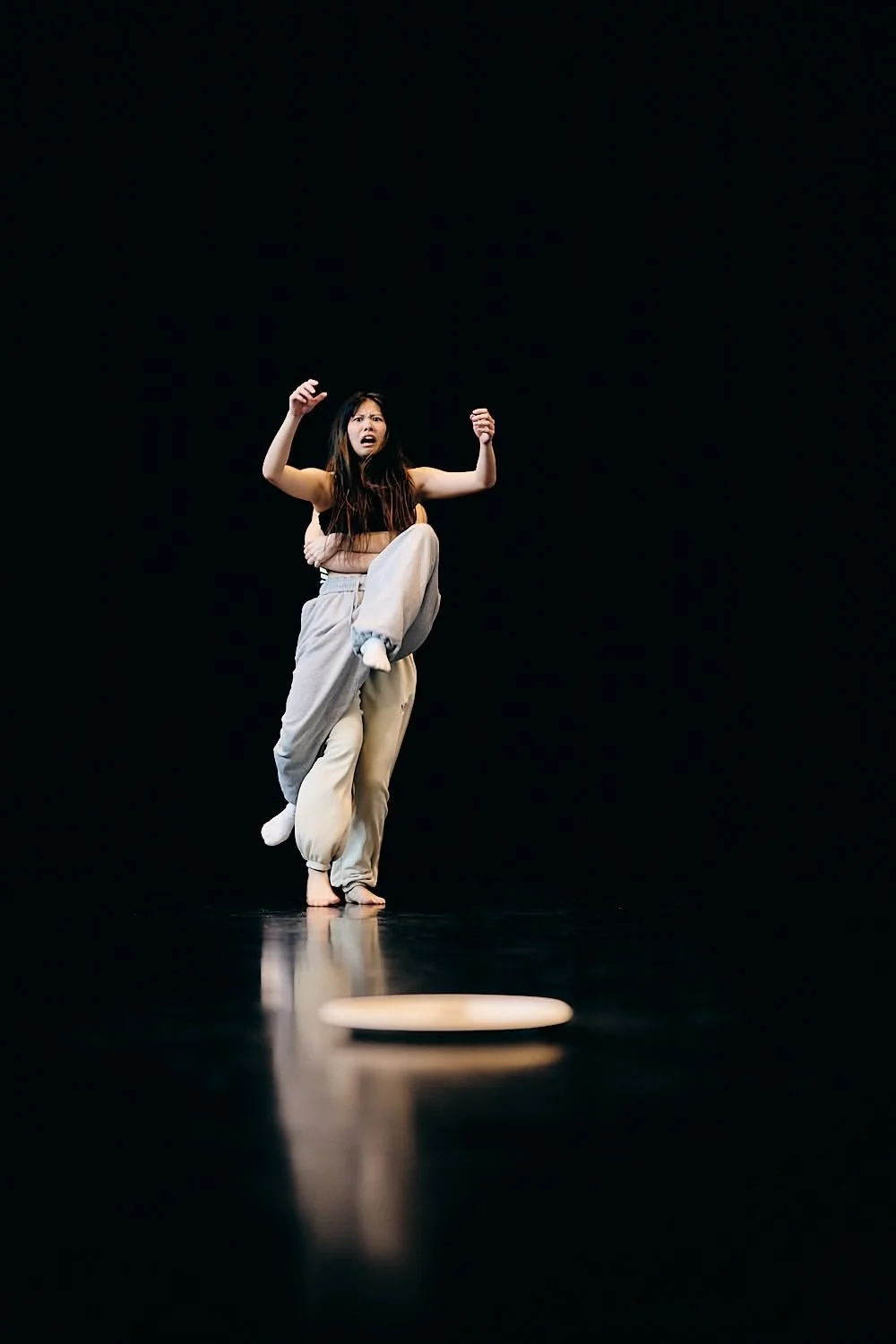 A woman performing on stage, wearing loose beige pants and a crop top, appears surprised or distressed, with her arms raised and one leg lifted. A ringed object or disc is on the dark stage floor in front of her.