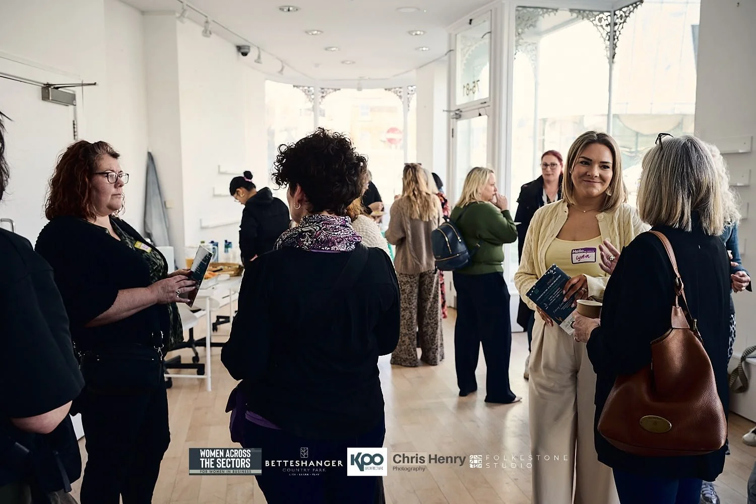 Group of women networking and conversing at an indoor event with large windows, some holding pamphlets or drinks, in a well-lit space with a wooden floor.