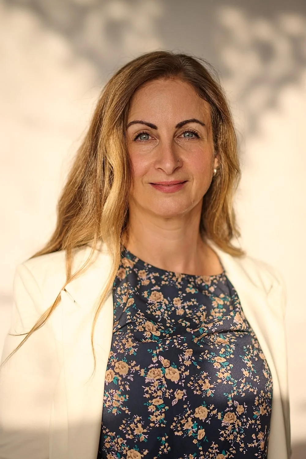 A woman with light skin, long wavy brown hair, and blue eyes wearing a floral top and a white blazer, smiling softly at the camera.