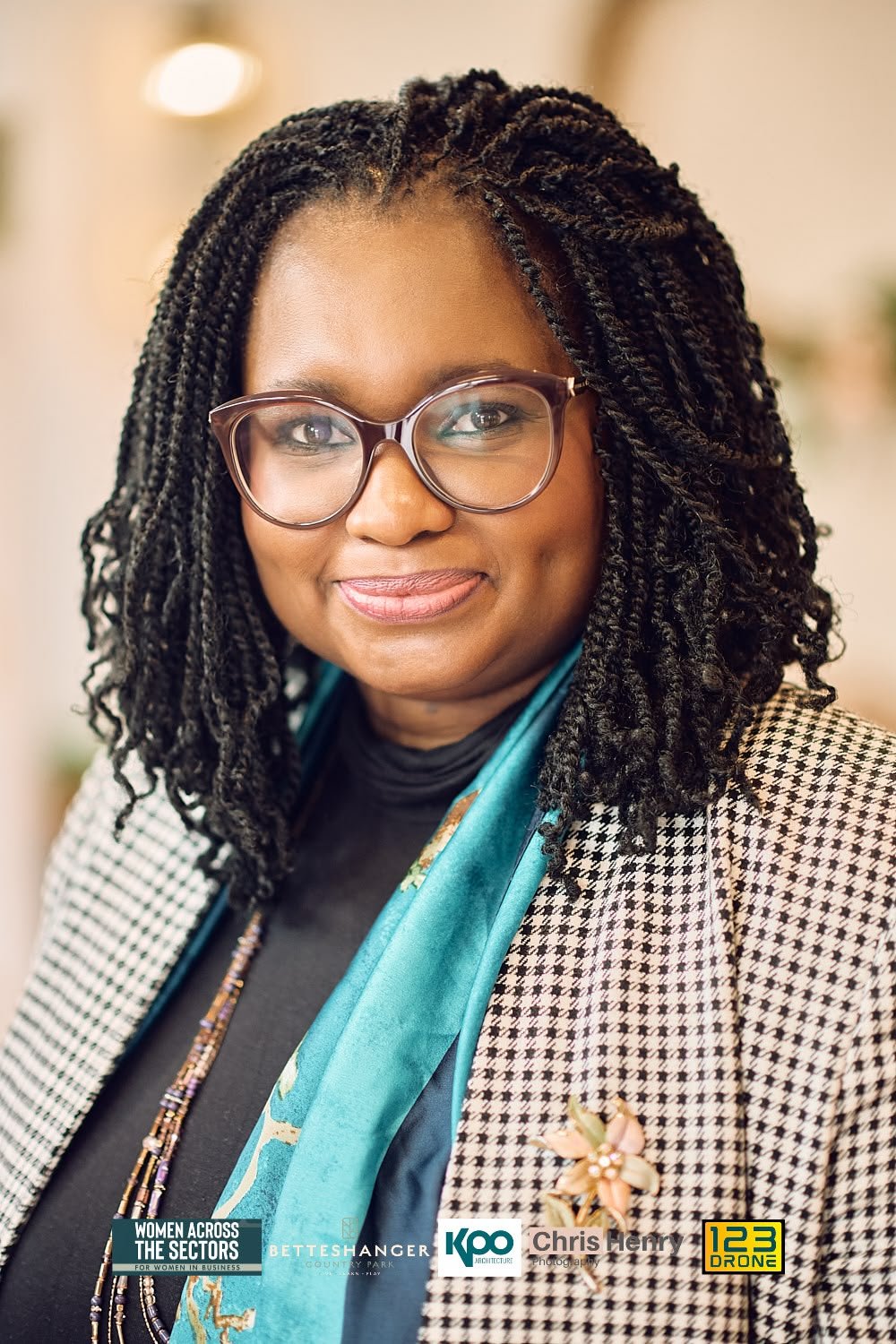A woman with glasses, dark curly hair in twists, and wearing a houndstooth blazer, black turtleneck, and a blue scarf, smiling at the camera.