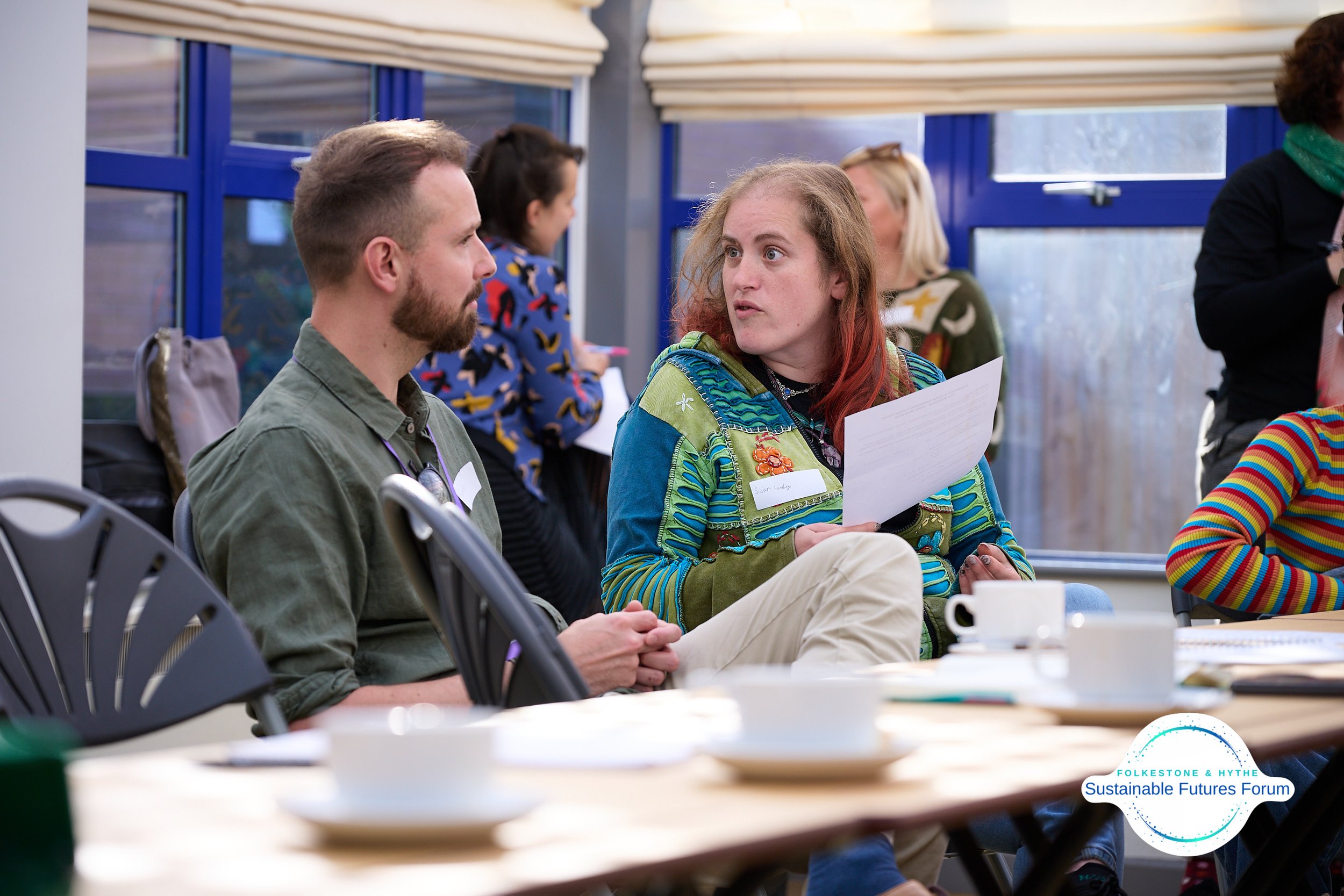 A woman with red hair talking to a man with a beard during a forum, with other attendees in the background and a logo for the Folkestone & Hythe Sustainable Futures Forum in the bottom right corner.