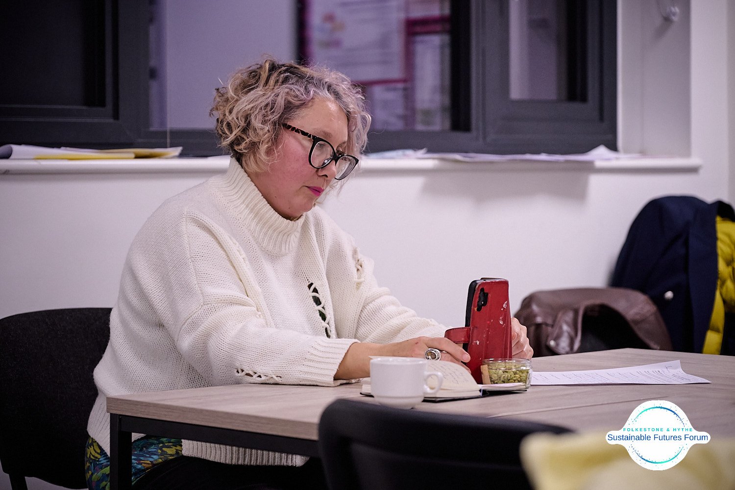 Woman with curly gray hair and glasses, wearing a white sweater, sitting at a table, looking at her phone. There is a notebook, a cup, and a small container of green nuts or seeds on the table. The background shows some papers and jackets.