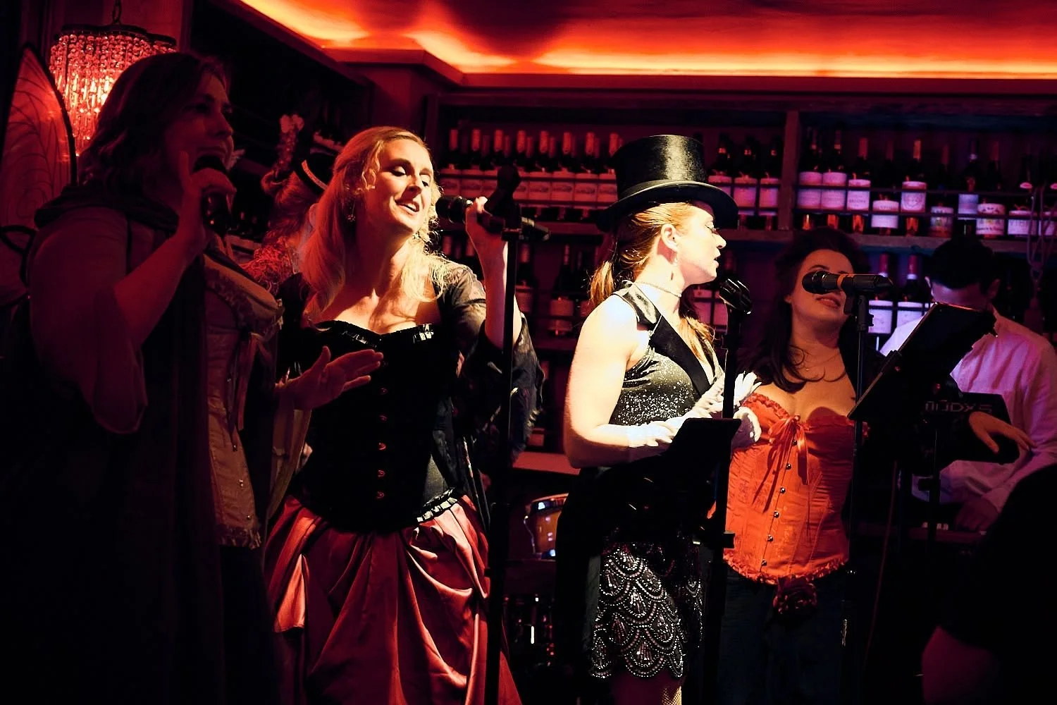 Group of women performing on stage in costume, singing into microphones, in a dimly lit bar or club with bottles on shelves in the background.