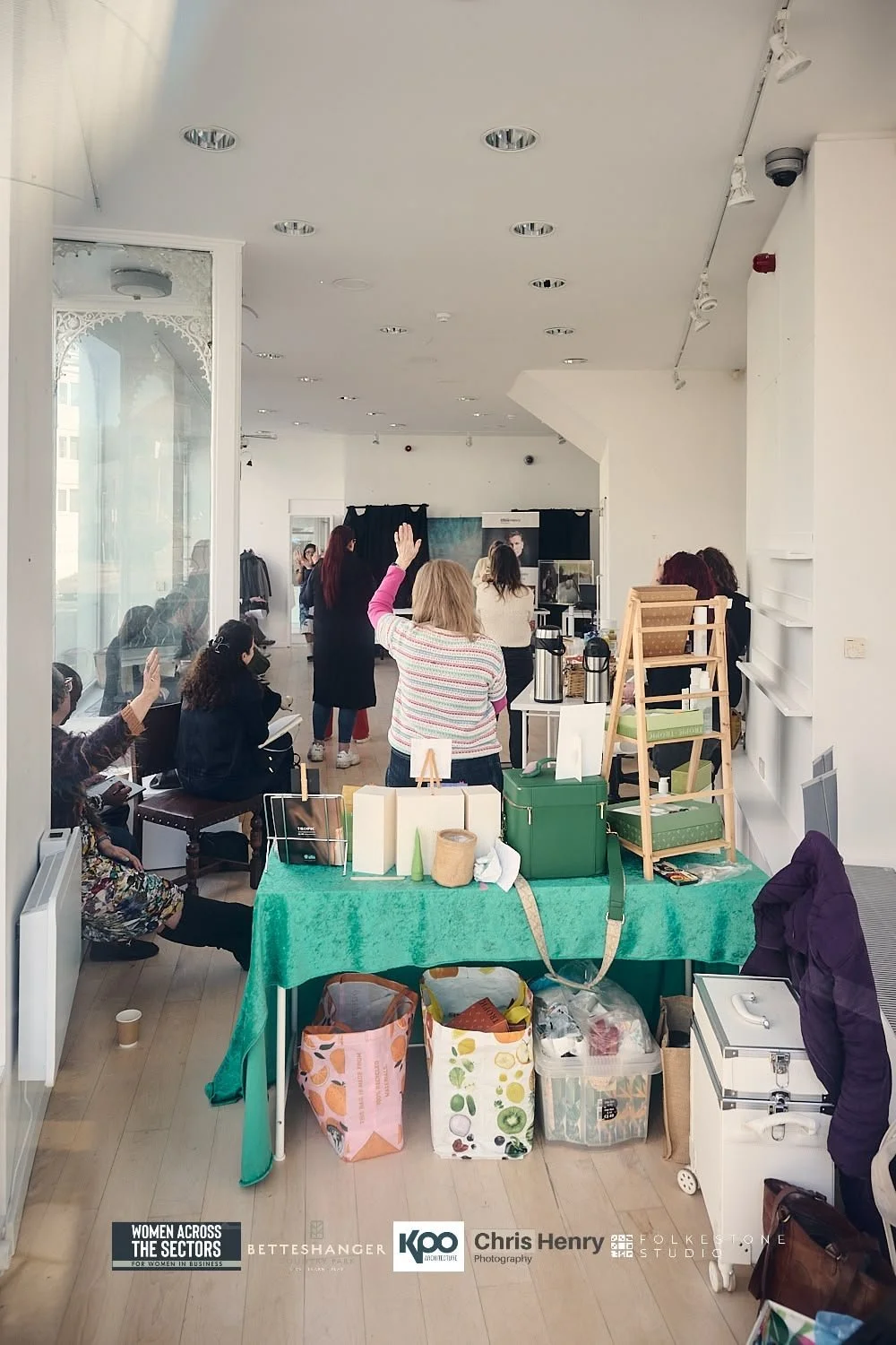 Women participating in a workshop or seminar in a bright, modern room. Some women are seated, while one woman stands with her hand raised. There is a table in the foreground with various items and supplies, including boxes, coffee dispensers, and bag