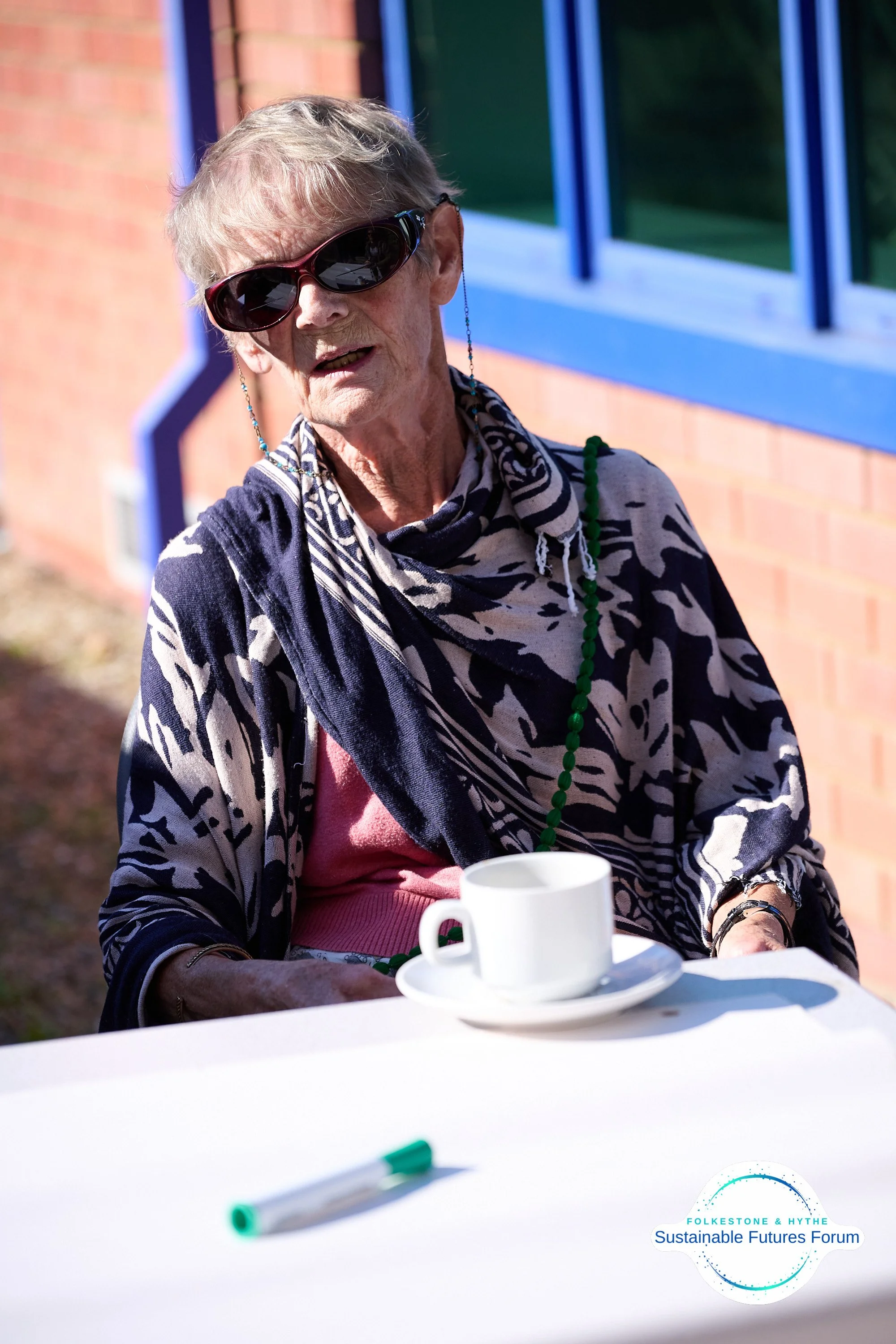 An elderly woman with short gray hair wearing sunglasses, a patterned scarf, a pink shirt, and a black and white jacket sitting outdoors at a table with a white cup and a green and white marker. There is a logo in the bottom right corner that reads '
