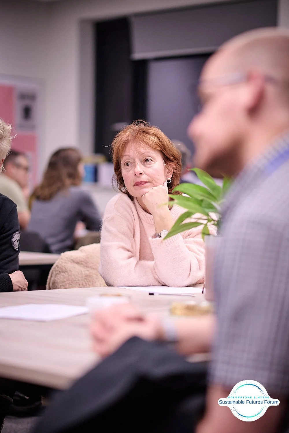 A woman with short red hair, wearing a pink sweater, is sitting at a table, resting her chin on her hand, listening during a meeting or discussion. In the foreground, there is a blurred man with glasses and a checkered shirt, and in the background, o
