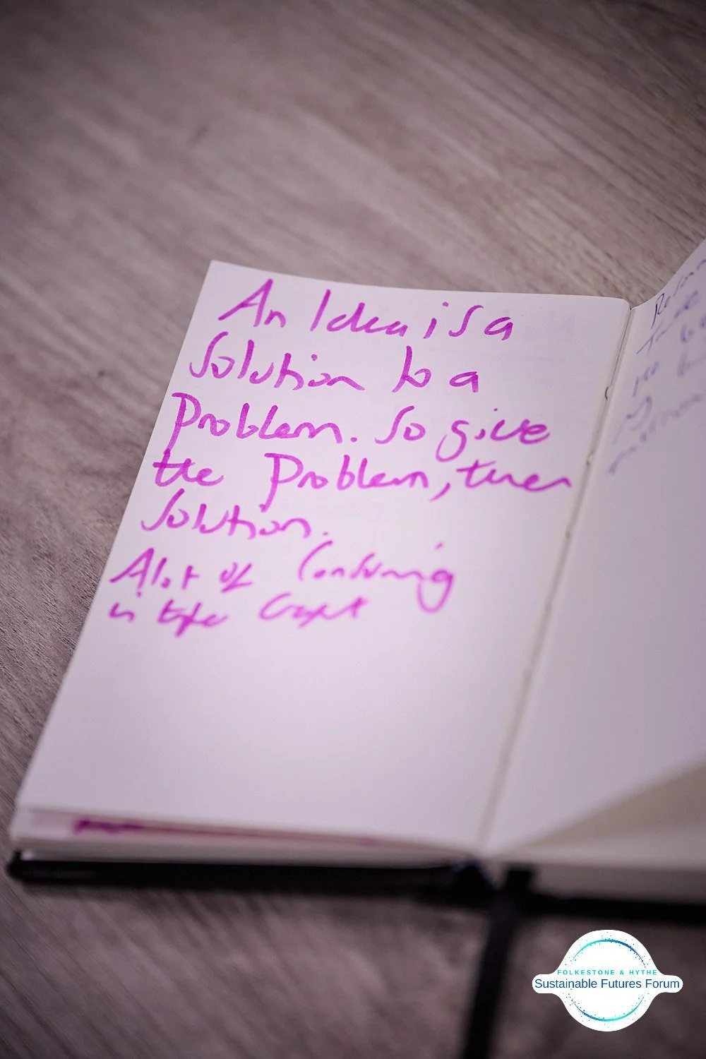 Open notebook with handwritten notes in pink ink, discussing problem-solving and analyzing costs, placed on a wooden surface with a logo of Folkestone & Aythe Sustainable Futures Forum in the bottom right corner.