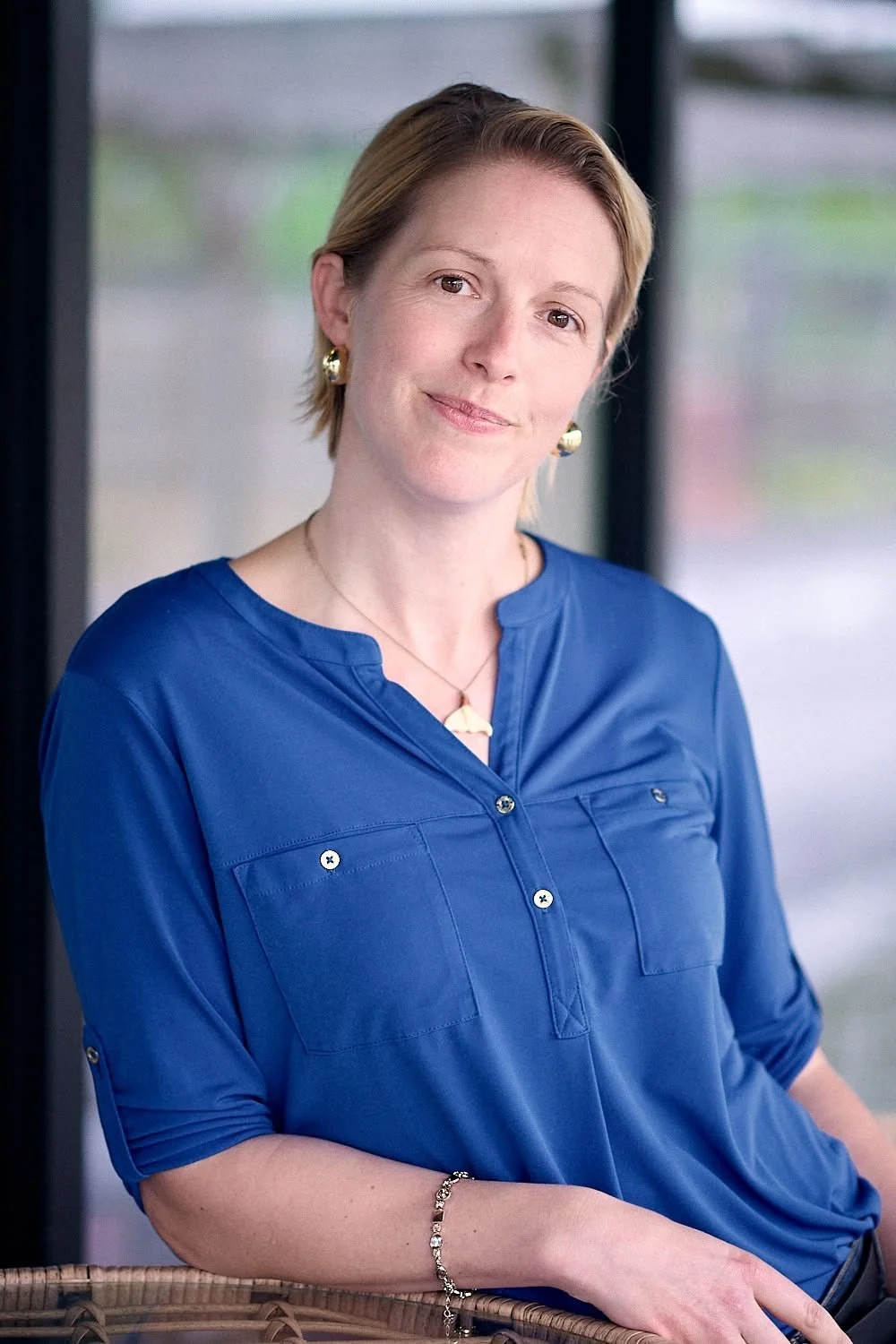 A woman with short blonde hair, wearing a blue blouse, gold earrings, and a delicate necklace, looks softly at the camera with a slight smile, sitting indoors near a window.